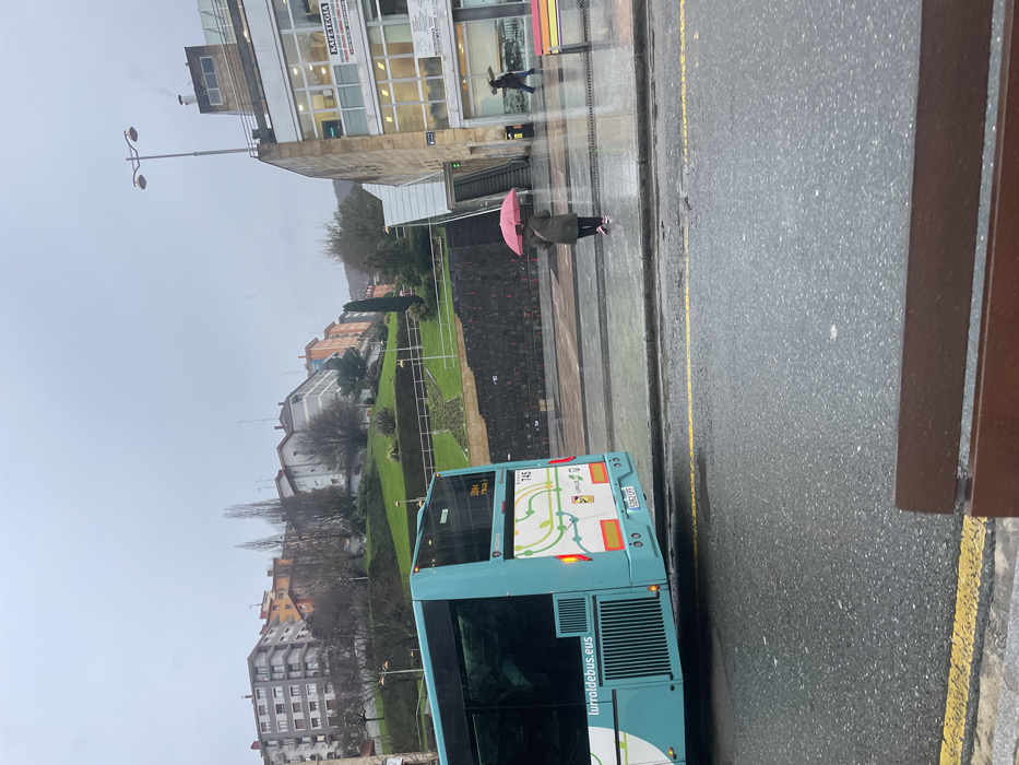 The image depicts an overcast and rainy urban street scene in Eibar, Spain. The perspective appears to be from inside a vehicle, looking outwards, with a brown frame partially visible on the right.
In the immediate foreground, a light blue and white public bus is positioned on a wet asphalt road. The rear of the bus faces away from the viewer, displaying the route number "145" and the web address "lurraldebus.eus". Its license plate, "6934 GPY", is also visible. Yellow painted lines mark the edge of the road and what appears to be a bus lane or parking area.
On the wet sidewalk next to the bus, a person dressed in a dark jacket is walking, holding a vibrant pink umbrella open, indicating the current or recent rainy weather. Further along the sidewalk, closer to a modern building with large glass windows, another individual is seen walking. This building features a sign with the word "KAFETEGIA" prominently displayed, along with other smaller, less legible text.
Across the street from the bus and sidewalk, a sloped, terraced green space or park is visible, featuring a distinct star-shaped garden bed. Beyond this green area, various multi-story residential buildings are nestled amongst mostly bare trees, suggesting a cooler season like winter or early spring. The sky is uniformly gray and cloudy, contributing to the muted lighting of the scene. A street lamp with two luminaires stands tall near the main building on the left side of the frame.