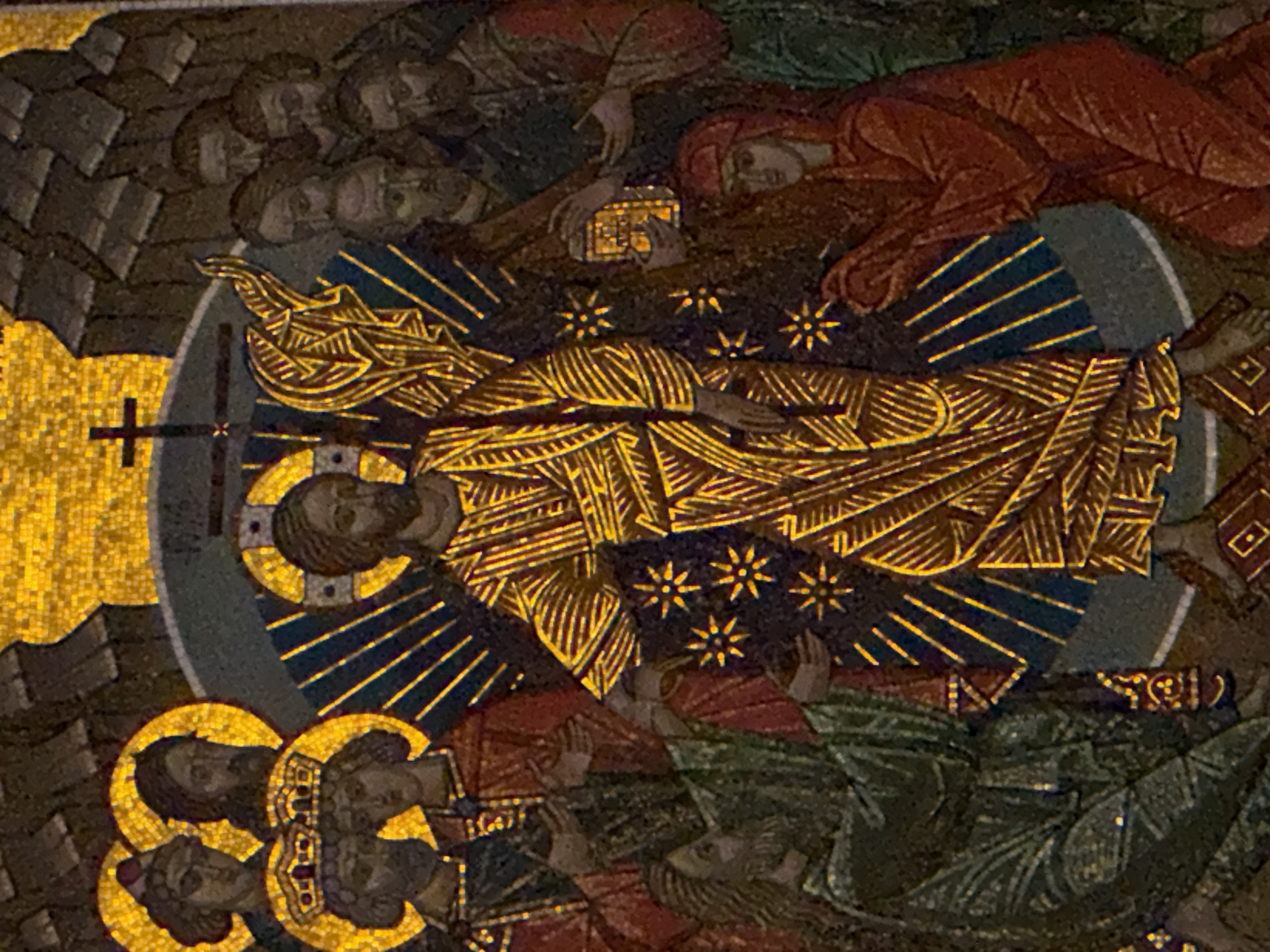 The image is a close-up of a mosaic depicting a religious scene, likely the Resurrection of Christ, in the Cathedral of Christ the Saviour in Moscow, Russia. The central figure, presumably Jesus, stands with arms outstretched, radiating golden light. He is adorned in a golden, striped robe. Surrounding him are figures who appear to be witnesses or disciples, some looking towards him with awe, others with bowed heads. One figure on the left is kneeling and wearing a green robe. Another figure on the right, dressed in red, is also kneeling and holding out an object that appears to be a container or lamp. The background is a mosaic of golden tesserae, with dark, rocky formations at the top. A cross is visible behind the central figure, and star-like motifs are scattered throughout the composition. The overall impression is one of divine triumph and reverence.