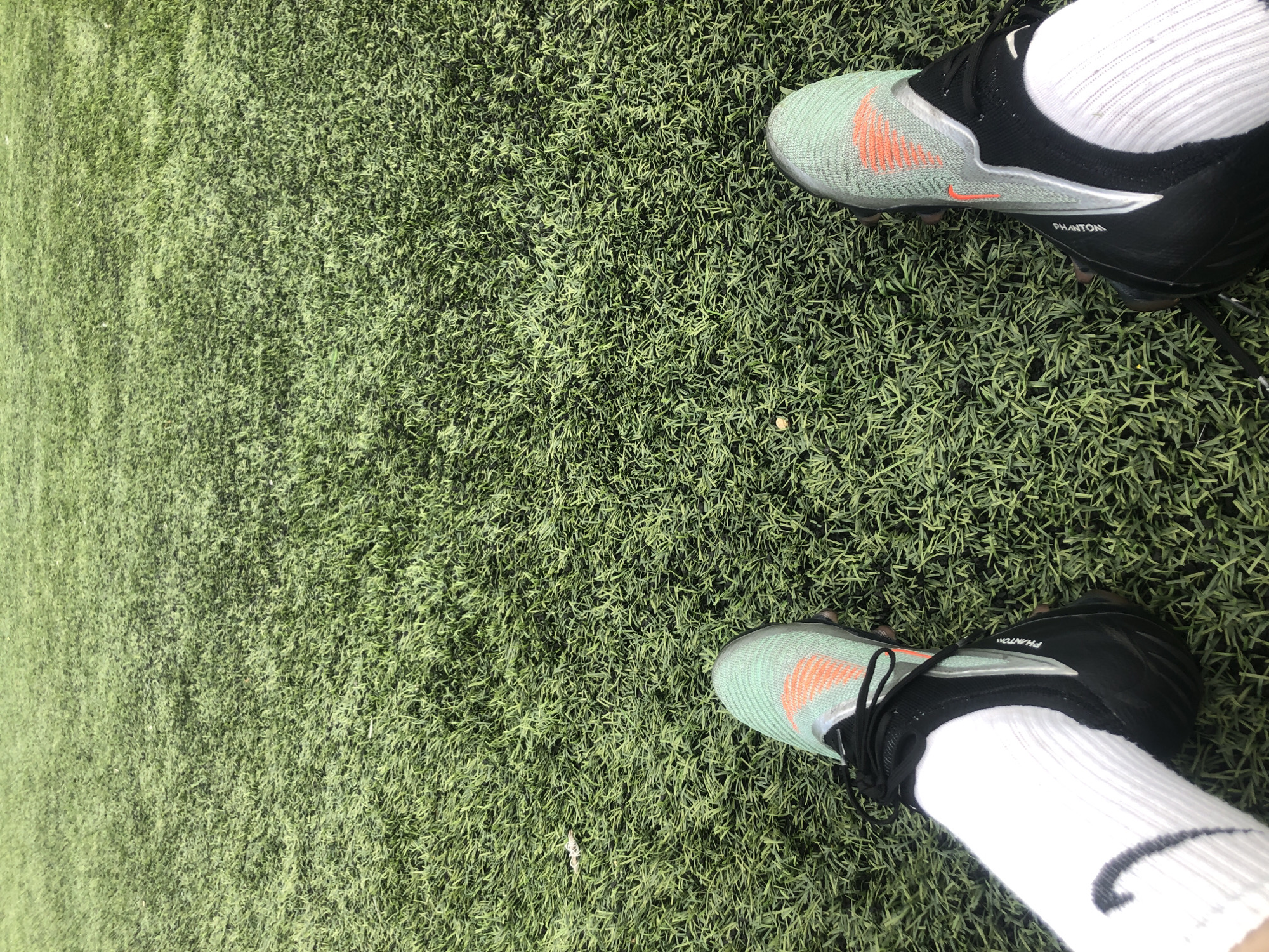 A person's feet are shown from a first-person perspective, resting on artificial turf. The person is wearing white socks with a black Nike swoosh logo visible on the left sock, and black soccer cleats with a light green and orange design. The cleats have the word "PHANTOM" printed on the side. The background is entirely composed of green artificial turf. There are no other people or objects clearly visible in the scene. The setting appears to be a sports field or park.