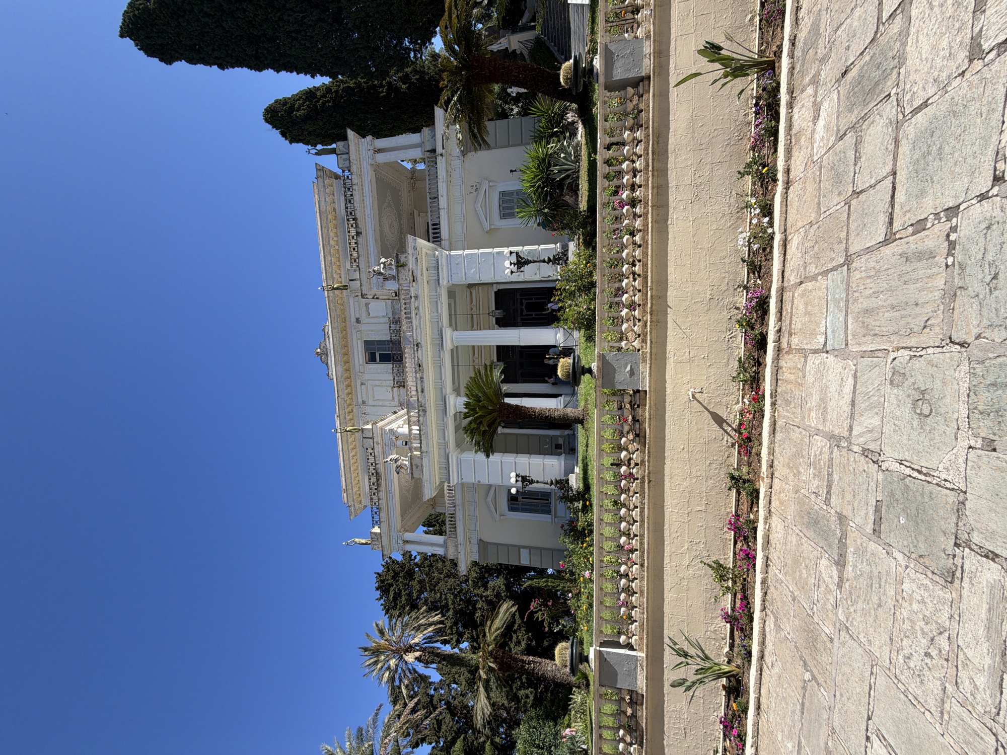 The image captures a grand, classical-style mansion or palace under a clear, bright blue sky, indicating a sunny day. The large, light-colored building, likely white or cream, features multiple stories, prominent columns at its main entrance, ornate balconies, and intricate detailing along its façades and roofline. Several large palm trees and tall, dark green coniferous trees, possibly cypresses, are visible around the property, suggesting a lush garden setting.
In the foreground, a paved pathway made of irregular stone blocks extends across the frame. Adjacent to this pathway is a low, ornate balustrade or fence, behind which lies a narrow garden bed vibrant with various flowering plants in shades of purple, pink, yellow, and white.
Notable architectural details include small, greenish statues or figures positioned on the building's upper balconies and roofline. Decorative street lamps are visible near the main entrance. On one of the upper façades, below the roofline, there is visible text that appears to be graffiti or a painted inscription, partially readable as "DEMEWEHE", possibly in a non-Latin script.
The overall scene conveys a sense of historical grandeur and peaceful beauty. No specific activities or people are distinctly visible, emphasizing the architectural and natural elements. The clear skies and strong light suggest a pleasant outdoor environment, consistent with the given location context of Gastouri, Greece, hinting at a Mediterranean or Neoclassical aesthetic.