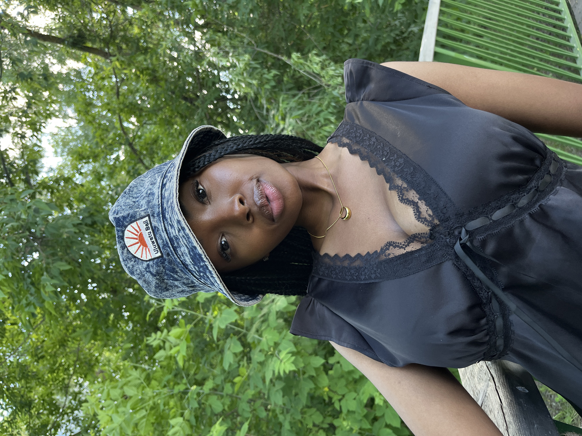 The media file shows a close-up, tilted portrait of a young Black woman outdoors. She is wearing a denim bucket hat with an emblem on the front that appears to depict a rising sun with Japanese text. Her hair is styled in braids that fall over her shoulders. She is wearing a black dress with lace trim along the neckline and straps, and a delicate gold necklace with a ring pendant.
The background is filled with lush green foliage, suggesting a natural setting with trees and other plants. Sunlight filters through the leaves, creating a dappled effect and indicating it is likely daytime. The woman is leaning on what appears to be a wooden railing or fence, possibly part of a park or outdoor seating area. She is looking towards the camera with a neutral to slight smile. The overall mood is relaxed and casual.
The visible text on the hat reads "Uguisu Ball," with the emblem showing a stylized red and orange sun. No other text is discernible. The location context of Kazan, Russia, is not directly evident from visual cues within the image itself, but suggests the scene takes place there. The weather appears to be pleasant and sunny, suitable for an outdoor activity.