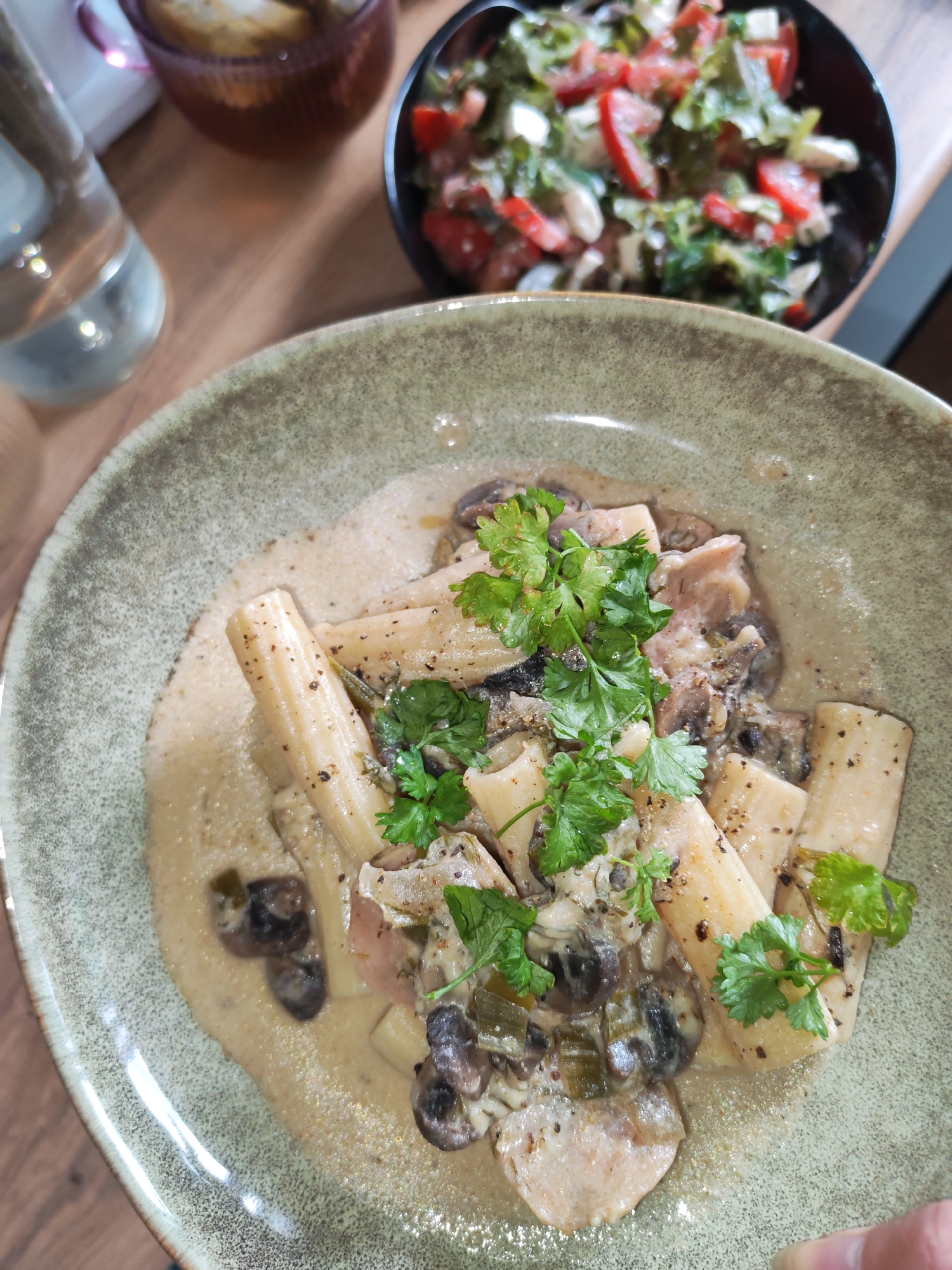 A close-up, overhead shot shows a plate of pasta with a creamy mushroom sauce. The pasta tubes are wide and short, and they are mixed with sliced mushrooms and bits of meat. Fresh parsley is sprinkled on top as a garnish. The plate is a textured, muted green color. In the background, out of focus, is a small bowl of salad with tomatoes and lettuce, and a glass of water. The setting appears to be a restaurant table.