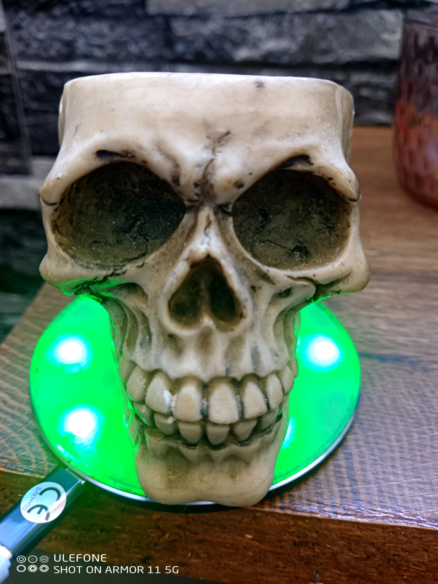 A frontal, slightly elevated perspective captures a decorative, off-white skull replica resting on a circular, brightly illuminated green LED light base. The skull, exhibiting aged or weathered dark accents within its eye sockets and nasal cavity, features a detailed set of upper teeth and an open top, suggesting its use as a container. The green light source below casts a distinct glow onto the lower portion of the skull. The skull and light base are positioned on a polished wooden surface with a visible grain. In the lower-left foreground, a black USB-C cable is partially visible, bearing a white label with a "CE" mark and the text "SN-HY01". The background includes a dark, textured wall constructed from block-like elements on the left, and a blurred, reddish-brown, textured object, possibly a vase or bottle, on the right. The image also contains a digital watermark in the bottom left corner, reading "Ulefone SHOT ON ARMOR 11 5G".