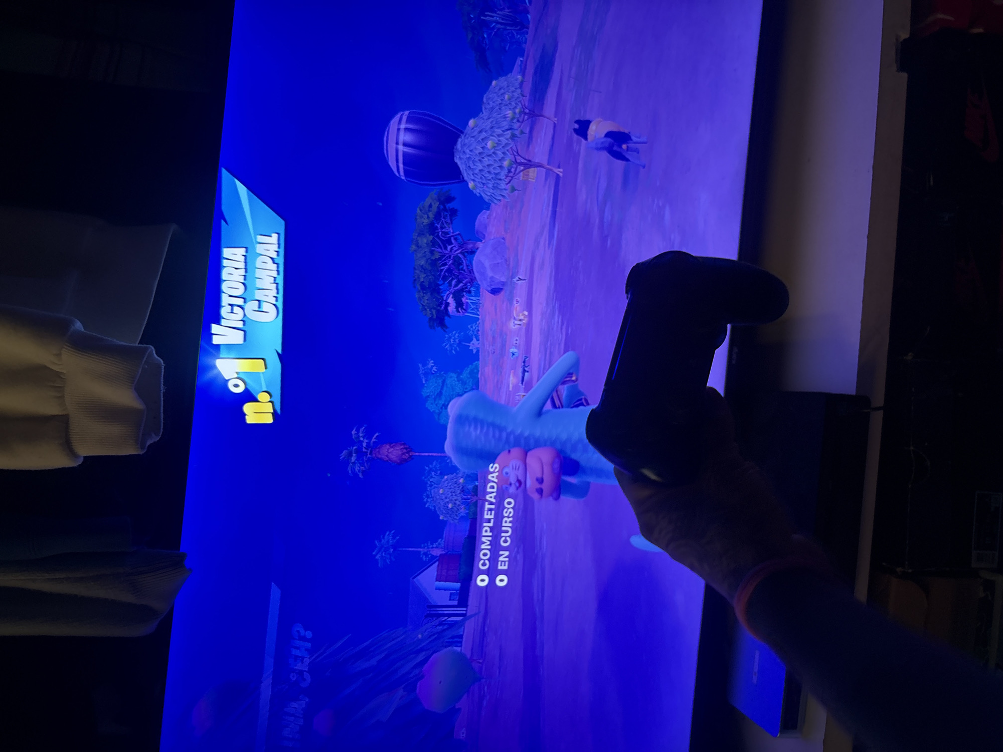 A person holds a black PlayStation controller in front of a television screen displaying a video game with the text "Victoria Campal." The game appears to be a brightly colored, animated environment with characters and objects. The game is in Spanish. The room is dimly lit, with clothing visible in the background.