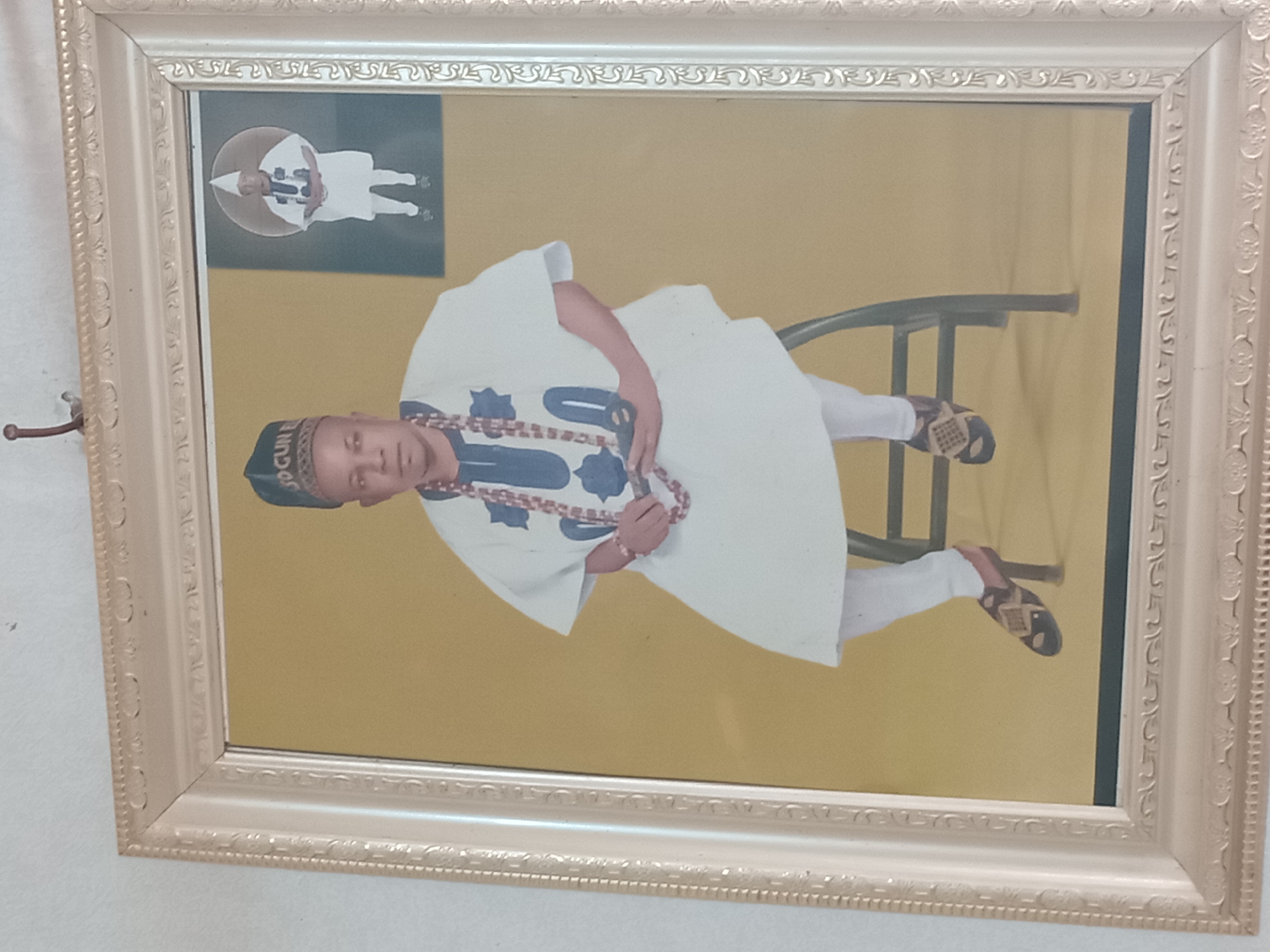 The image shows a framed photograph hanging on a wall. The photograph depicts a man seated on a chair. He wears a white outfit with blue and black accents, and a beaded necklace. He has a dark blue cap on his head with a word on it. In the upper left corner of the photograph is another image of a man in white against a dark gray background. The picture frame is ornate and off-white. The setting appears to be indoors. Location identified is the city of Shagamu, Nigeria.