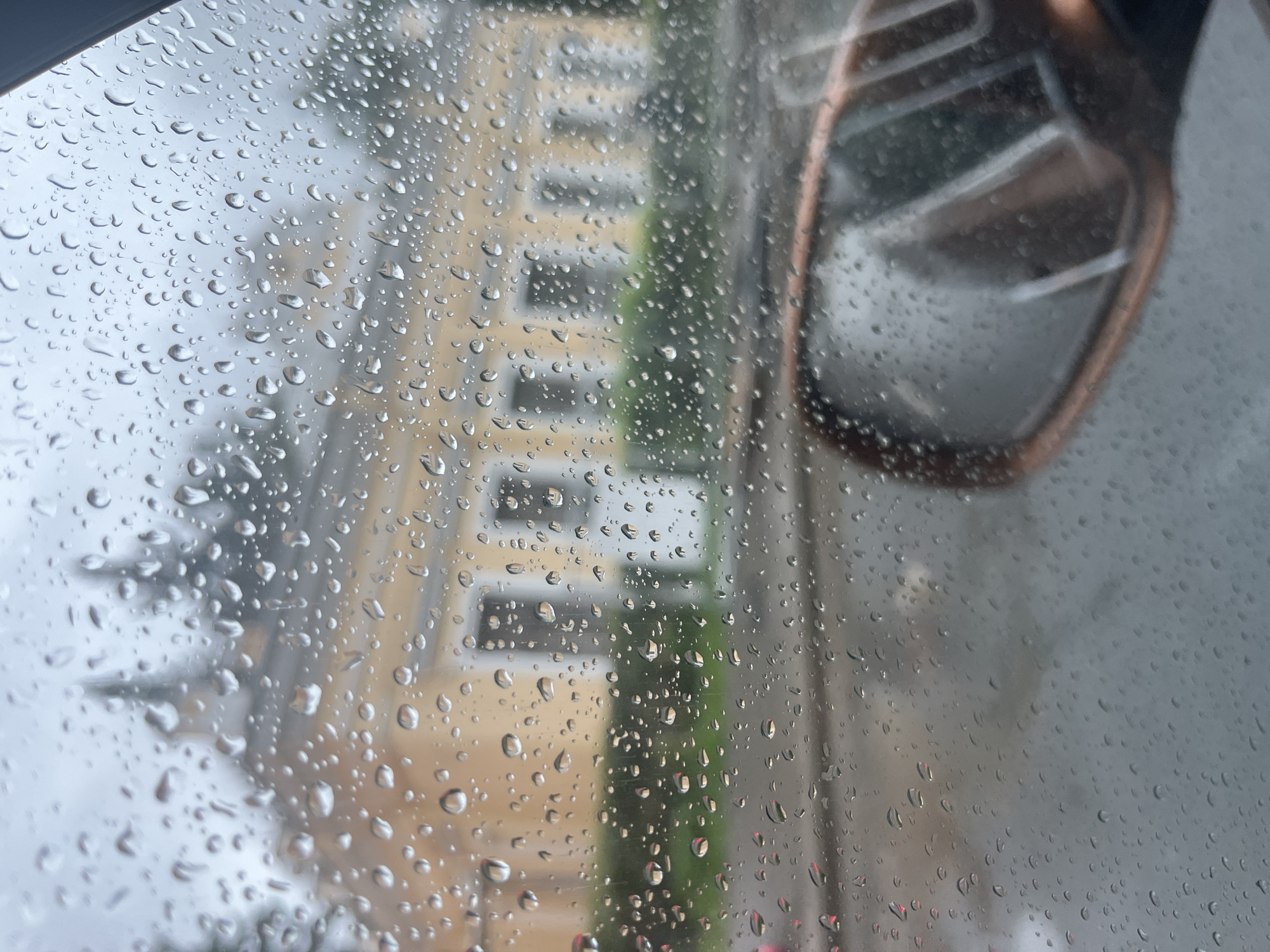 The image is a close-up shot of raindrops on a car window, looking out onto a street scene in Saint Petersburg, Russia. A yellow building with multiple windows and a pointed turret is visible in the background, along with greenery. The reflection of a car's side mirror, with a rose gold frame, is in the lower right portion of the image. The sky appears overcast, consistent with rainy weather. The overall impression is one of a wet, cloudy day in a European city.