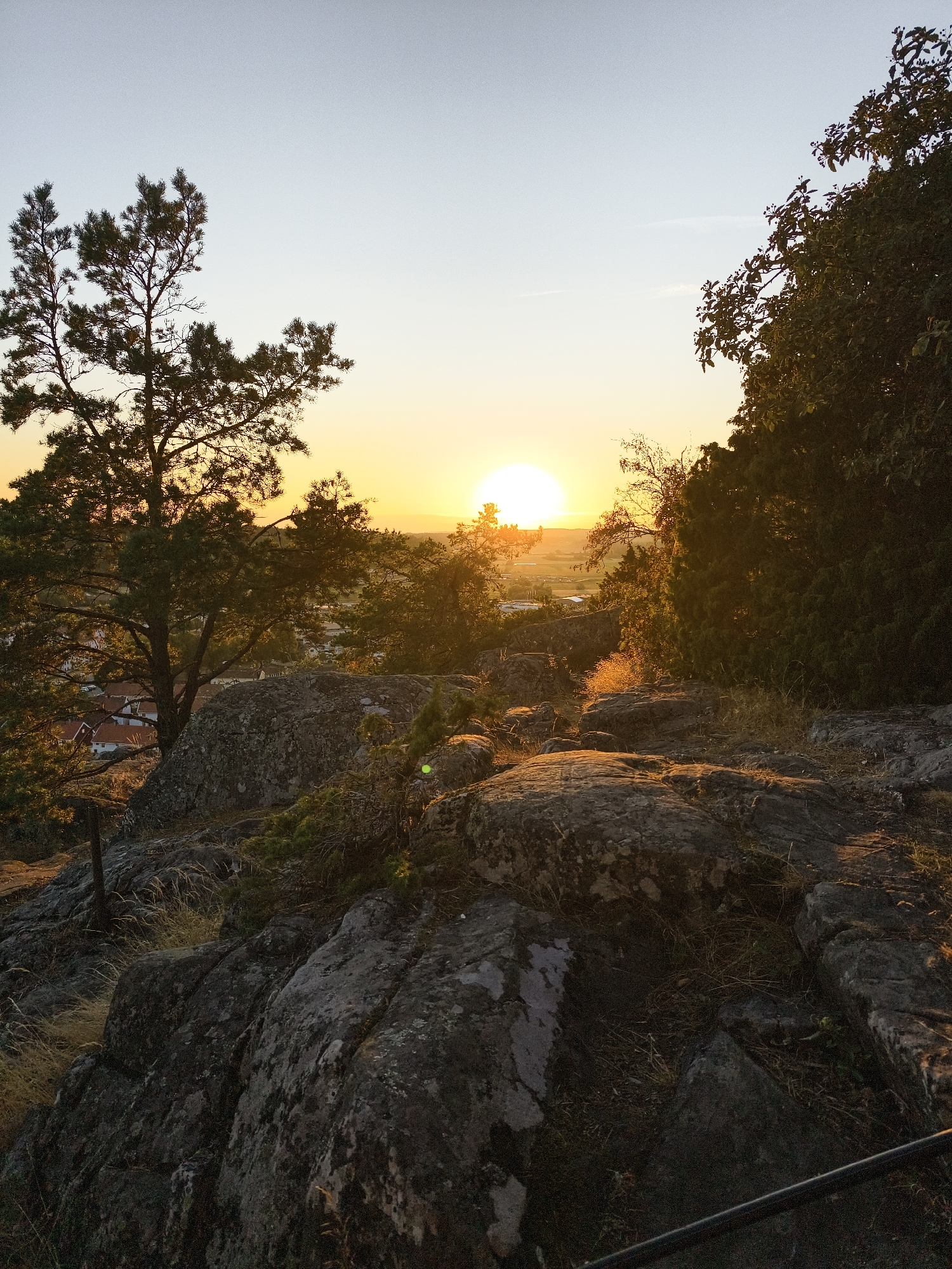 The image depicts a natural landscape at sunset in Söderköping, Sweden. The foreground is dominated by large, weathered rocks covered in lichen and scattered with dry grass. A dark metal railing runs along the bottom right edge of the frame. Behind the rocks, a cluster of trees, including a tall pine, are silhouetted against the bright sky. In the middle ground, a small town with red-roofed houses is visible amongst further trees. The sun, a bright orange orb, is setting on the horizon, casting a warm glow over the scene and creating a gradient of yellow and orange in the sky. The overall impression is one of tranquility and natural beauty as daylight fades.