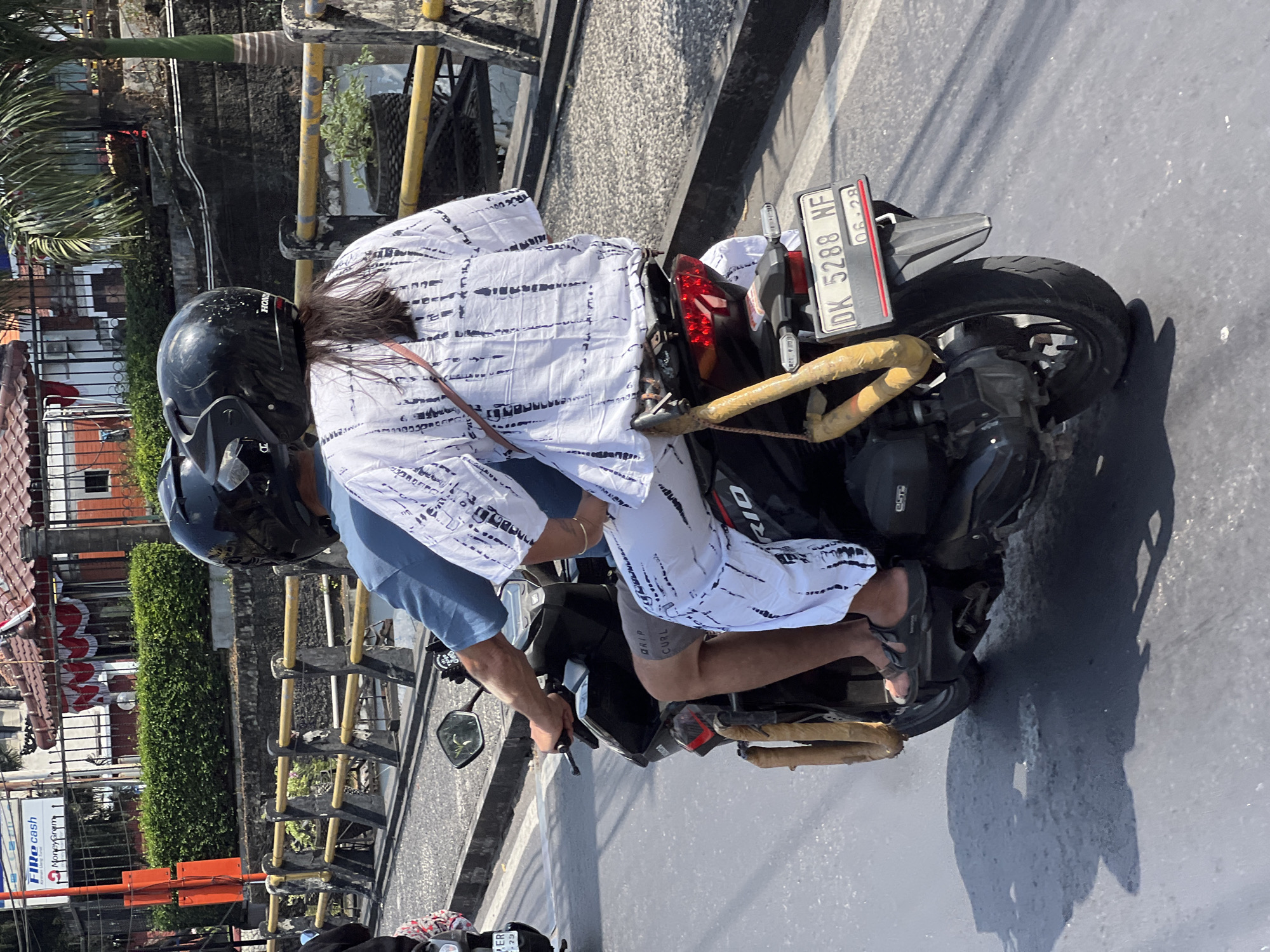 Two individuals, both wearing black helmets, ride on a black scooter along a paved road. The person in front, presumably the driver, wears a blue t-shirt and grey shorts. The passenger in the rear wears a white, loose-fitting top with black tie-dye patterns and white pants with similar black patterns. The scooter has a license plate displaying "DK 5288 NF" and a sticker with "06-28". The scooter is equipped with yellow protective padding on the sides, wrapping around a metal bar. In the background, a black metal fence with yellow and grey posts runs alongside the road, separating it from a lush green hedge. Behind the hedge, a building with a red tiled roof and white decorative elements is visible. The scene appears to be in Denpasar, Indonesia, given the license plate format and the general environment. Sunlight casts shadows on the road, indicating a bright, sunny day.