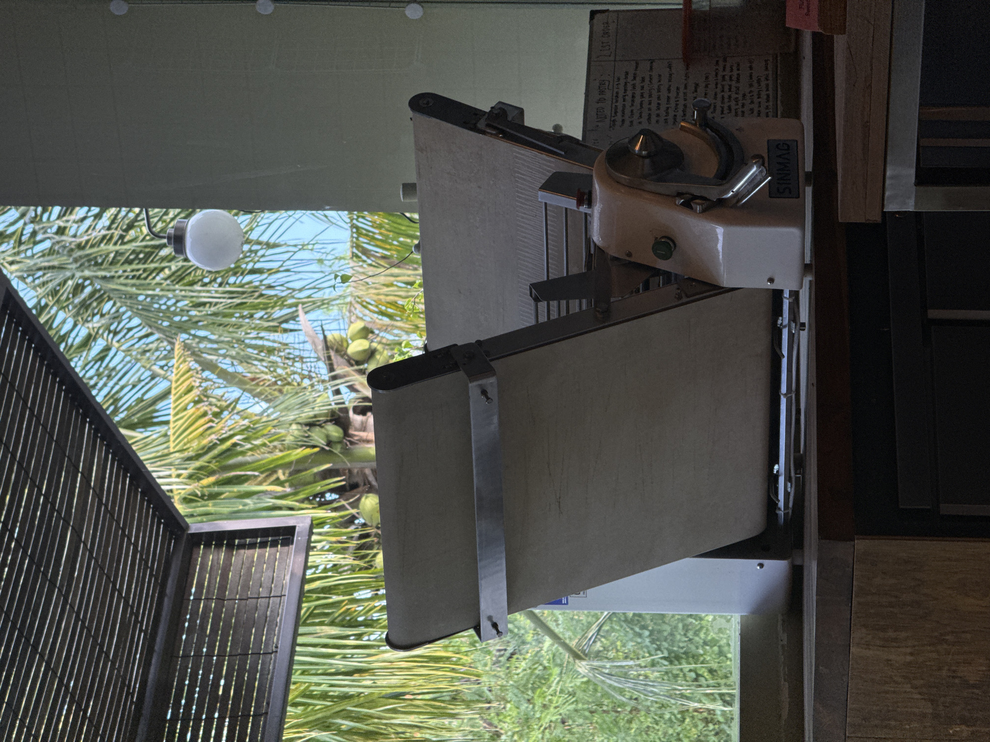 The image presents an indoor view looking out onto a bright tropical setting. In the foreground, a cream-colored industrial machine, identified by the brand "SINMAG" on its side, is prominently displayed. This machine, likely a pastry or bread slicer, features various metal components, a lever, and a green button. Behind it, a wooden counter or surface is visible. Above and behind the machine, a piece of paper or board is taped up, displaying handwritten notes and text including "NOTED TO PASTRY" and "LIST ORDER." Other partially legible words like "soon loss no moon Re," "M.S," "Flu," and "Sweet" are also present, suggesting order details or instructions.
Through a large window or opening, a vibrant outdoor scene unfolds. Lush green palm trees, some bearing visible coconuts, dominate the exterior landscape under a clear, bright blue sky. This indicates a sunny day in a tropical environment, consistent with the North Kuta, Indonesia location. A white spherical light fixture hangs from the ceiling in the indoor space, partially intersecting the view of the outdoor foliage. To the left, a dark slatted structure, possibly a screen or partition, frames the interior perspective. No individuals are present, and no specific activity is underway; the scene depicts a static setup, likely within a bakery, cafe, or kitchen.