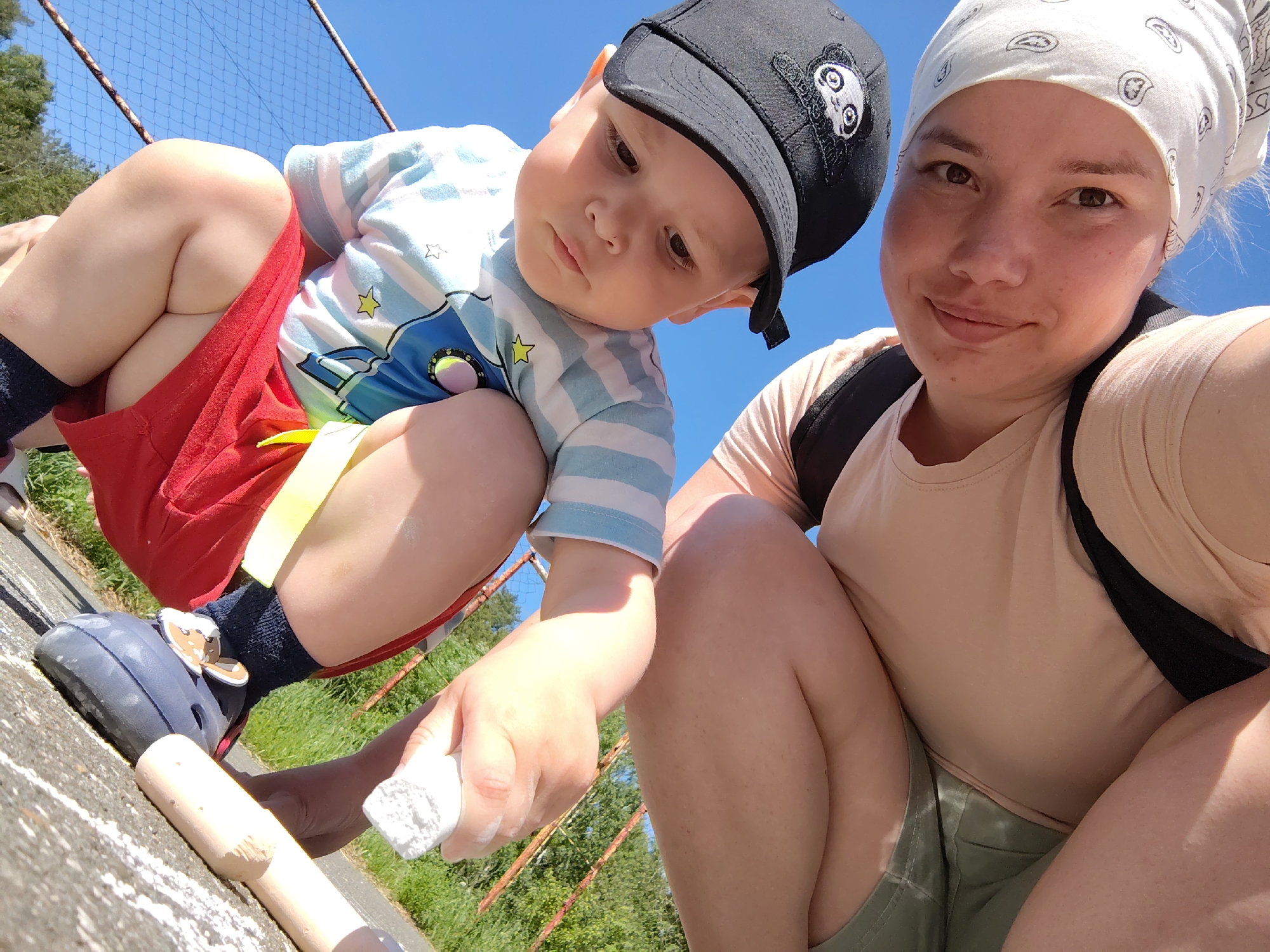 A woman and a toddler squat on a light gray surface in the city of Markovskii, Russia. The woman has fair skin, brown eyes, and a slight smile. She wears a white paisley bandana, a light pink t-shirt, and olive green shorts. A black backpack strap is visible on her shoulder.
The toddler, who is fair-skinned, wears a black baseball cap with a panda cartoon design, a blue and white striped shirt with cartoon images, red shorts, a yellow tag, dark blue socks, and dark blue Crocs with a bear face decoration.
Both the woman and the toddler are holding white chalk. There are other pieces of chalk on the surface. A chain-link fence and greenery are visible in the background under a clear blue sky.