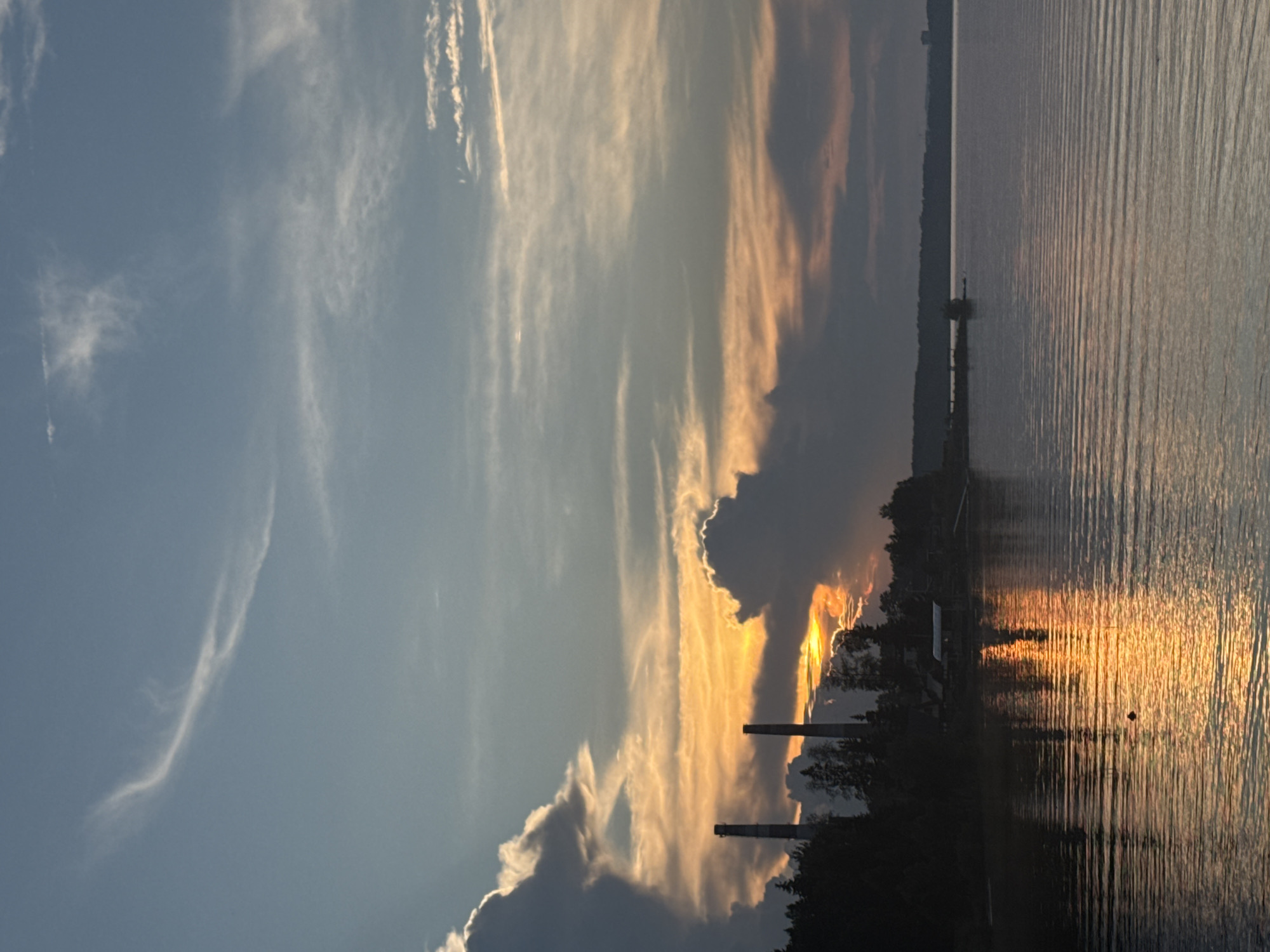 The image depicts a sunset over the Kama River in Chaikovsky, Russia. In the foreground, the river's surface is rippled, reflecting the warm hues of the setting sun. A small, dark object, possibly a bird, is visible on the water. The middle ground shows a tree-lined shore with two tall, industrial smokestacks silhouetted against the sky. A few buildings are nestled amongst the trees. The sky is a blend of blues and oranges, with scattered clouds catching the last rays of sunlight. In the distance, the far bank of the river is visible as a dark, undulating line, with a few indistinct structures on the horizon. The overall atmosphere is serene and highlights the industrial and natural elements of the Chaikovsky landscape.