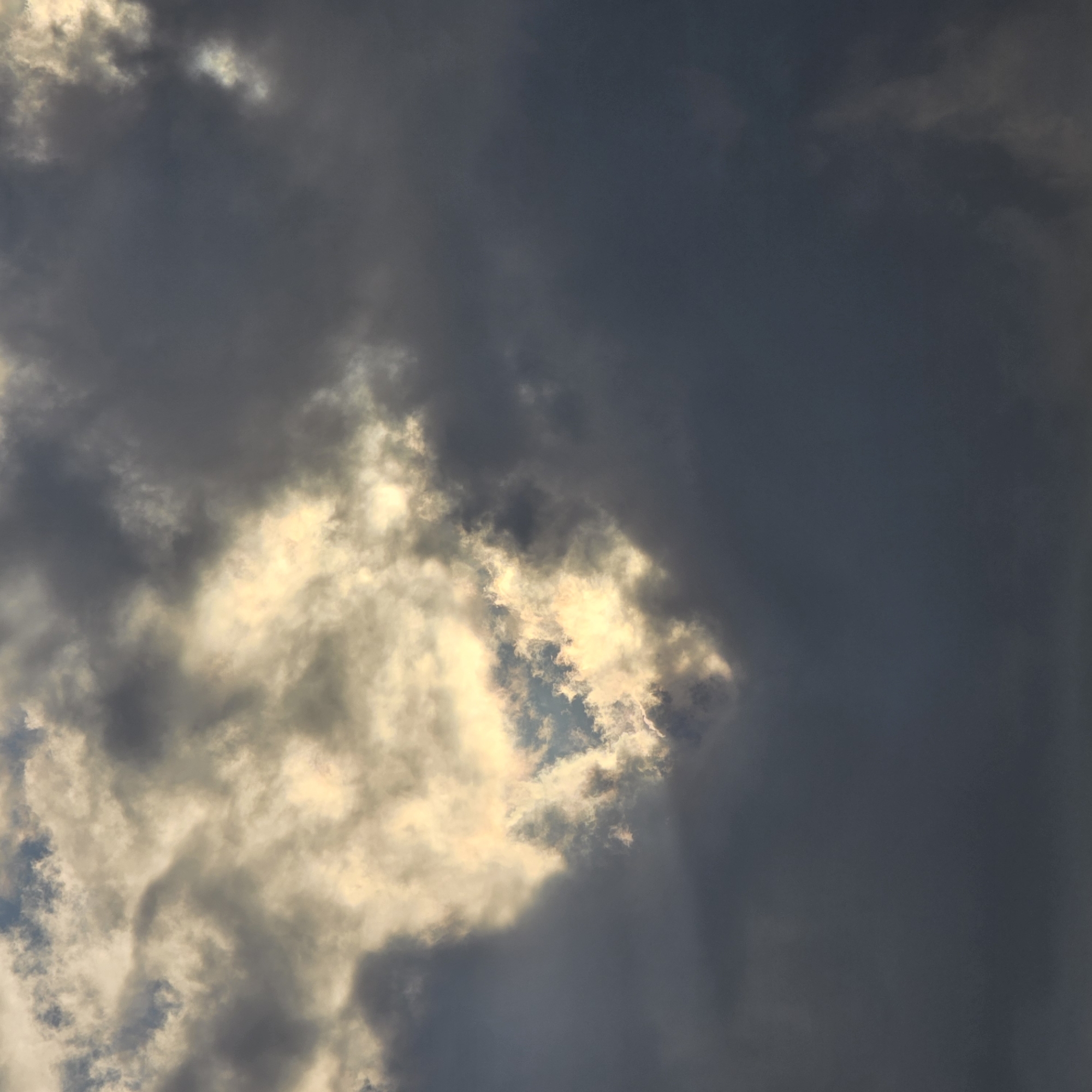 The image is a close-up of a cloudy sky, likely taken from a ground-level perspective in Belokurikha, Russia. The clouds are a mix of dark, heavy storm clouds and lighter, brighter clouds illuminated by sunlight peeking through. There appears to be a gap or opening in the clouds where the sun's rays are intensely shining through, creating a dramatic contrast. The lighting suggests it is either early morning or late afternoon. There are no people, animals, or man-made structures visible in the frame, focusing solely on the atmospheric conditions of the sky.