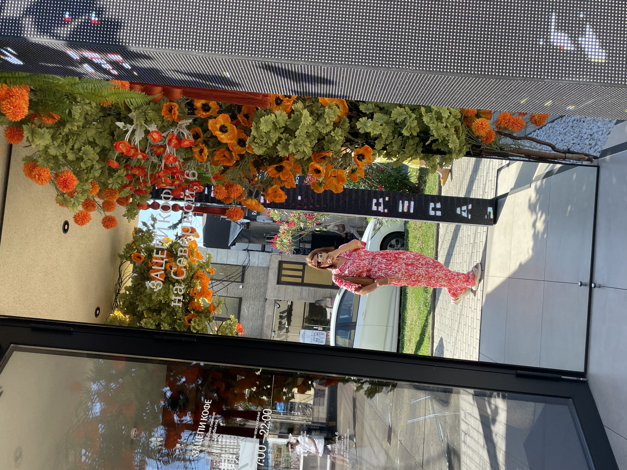 A woman in a pink floral dress stands in front of a building facade, taking a picture with her phone. She is positioned to the right of a large glass storefront with text reflected on it that reads "ЗАЦЕПИ КОФЕ" and "КОФЕЙНЯ ЛИЧНЫХ СОБЫТИЙ" and "7:00-22:00." The storefront has an entrance that appears to be open. To the right of the storefront is a decorative floral arrangement featuring bright orange and green foliage. The floral arrangement is positioned in front of a large, dark, textured wall with a grid pattern, possibly a digital display. In the background, a white car is parked on a paved area, and buildings with windows and balconies are visible. The scene appears to be on a sunny day, with shadows cast on the ground. The text on the storefront and the floral decorations suggest a commercial establishment, likely a coffee shop. The presence of buildings and vehicles indicates an urban environment. The provided information specifies the location as Sochi, Russia.