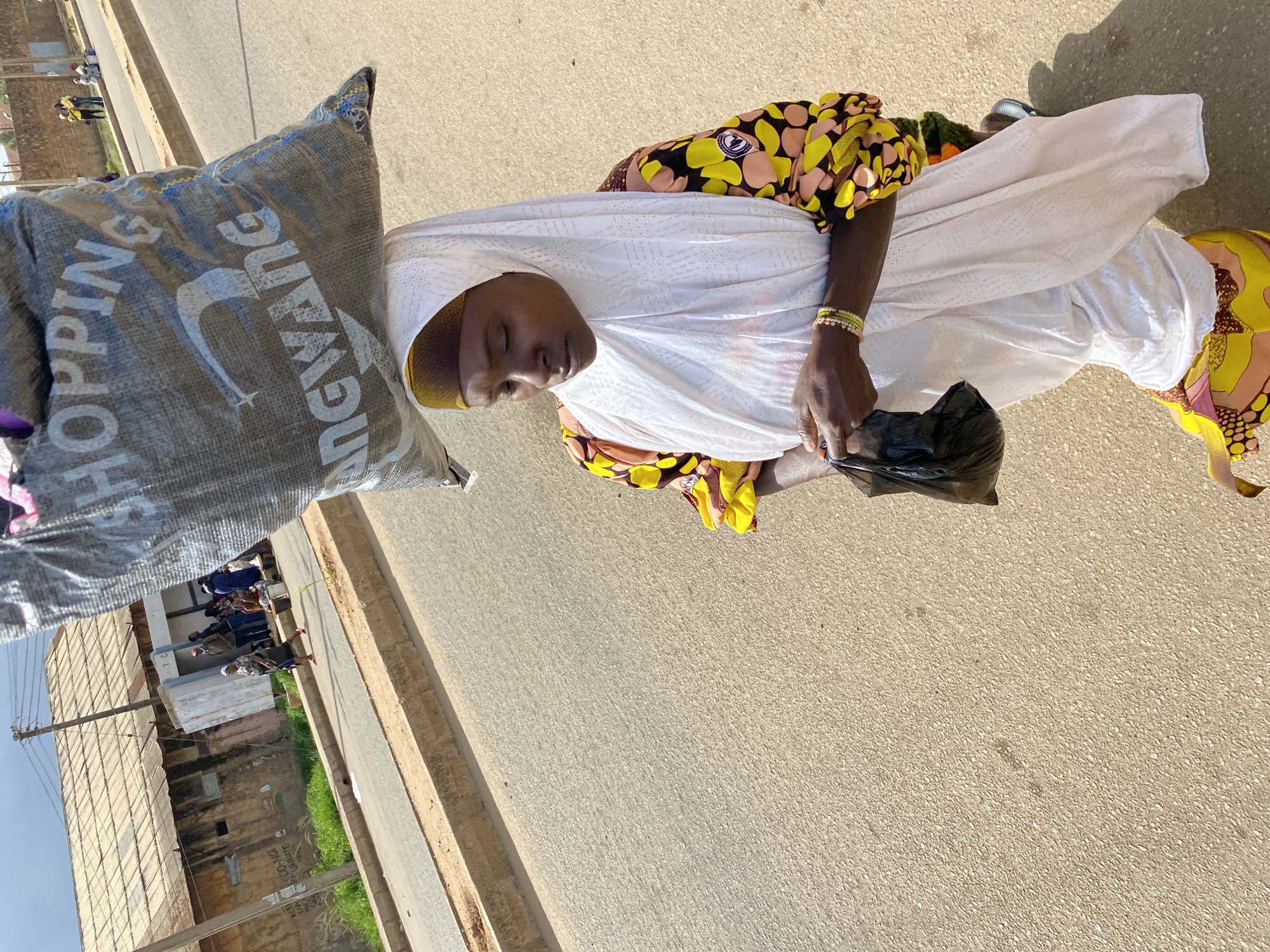A woman, wearing a white hijab and a brightly colored African print dress, walks down a paved street. She carries a large, grey shopping bag on her head. The bag has white lettering that reads "SHOPPING" and "ANCIENT WANG". In her right hand, she clutches a black plastic bag. The street is lined with a concrete curb and a dusty shoulder. In the background, buildings with corrugated metal roofs and a power pole are visible. Several other people are seen in the distance, some standing and others walking. The scene appears to be in an urban environment in Bukuru, Nigeria. The lighting suggests it is daytime with bright sunlight.