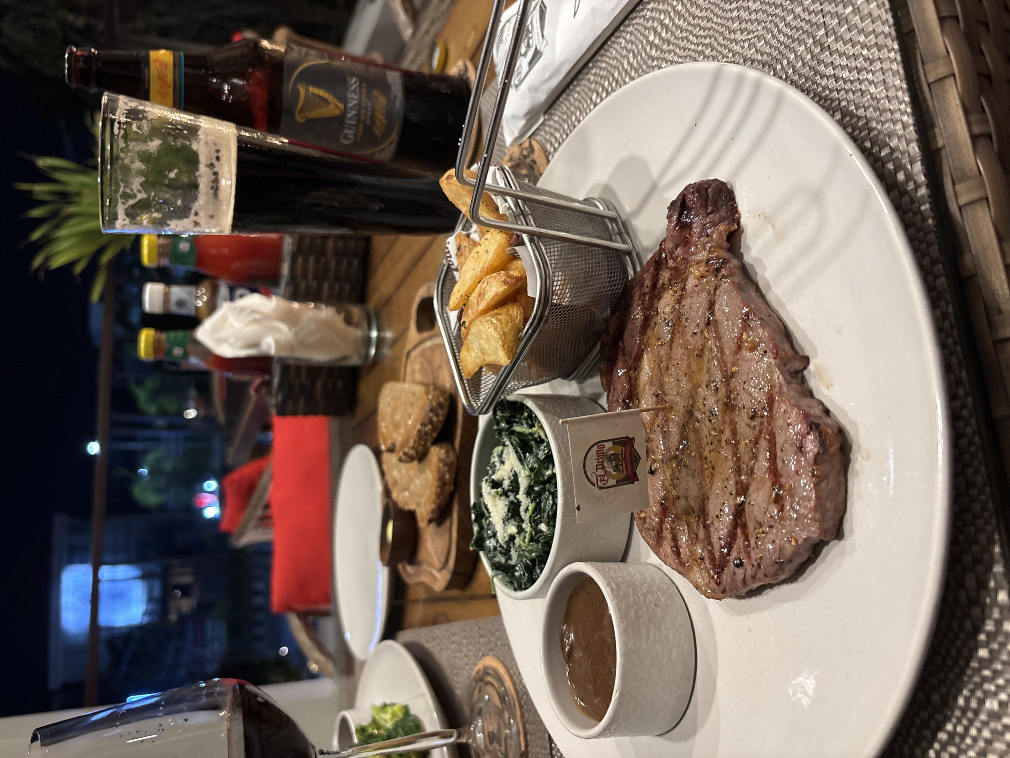 The image shows a table setting in a restaurant, likely outdoors, in Kuta Selatan, Indonesia, during the evening. On the table, there is a white plate with a grilled steak, served with a small basket of fries, a small dish of spinach with cheese, and a small bowl of sauce. A flag with the words "El Primo" is inserted into the steak. To the left of the plate, there are bread slices and a small dish, and another plate with green vegetables and a wine glass are partially visible. A glass of Guinness is placed behind the plate, along with a bottle of Guinness and other unidentified bottles. In the background, city lights are visible. The table is covered with a woven placemat.