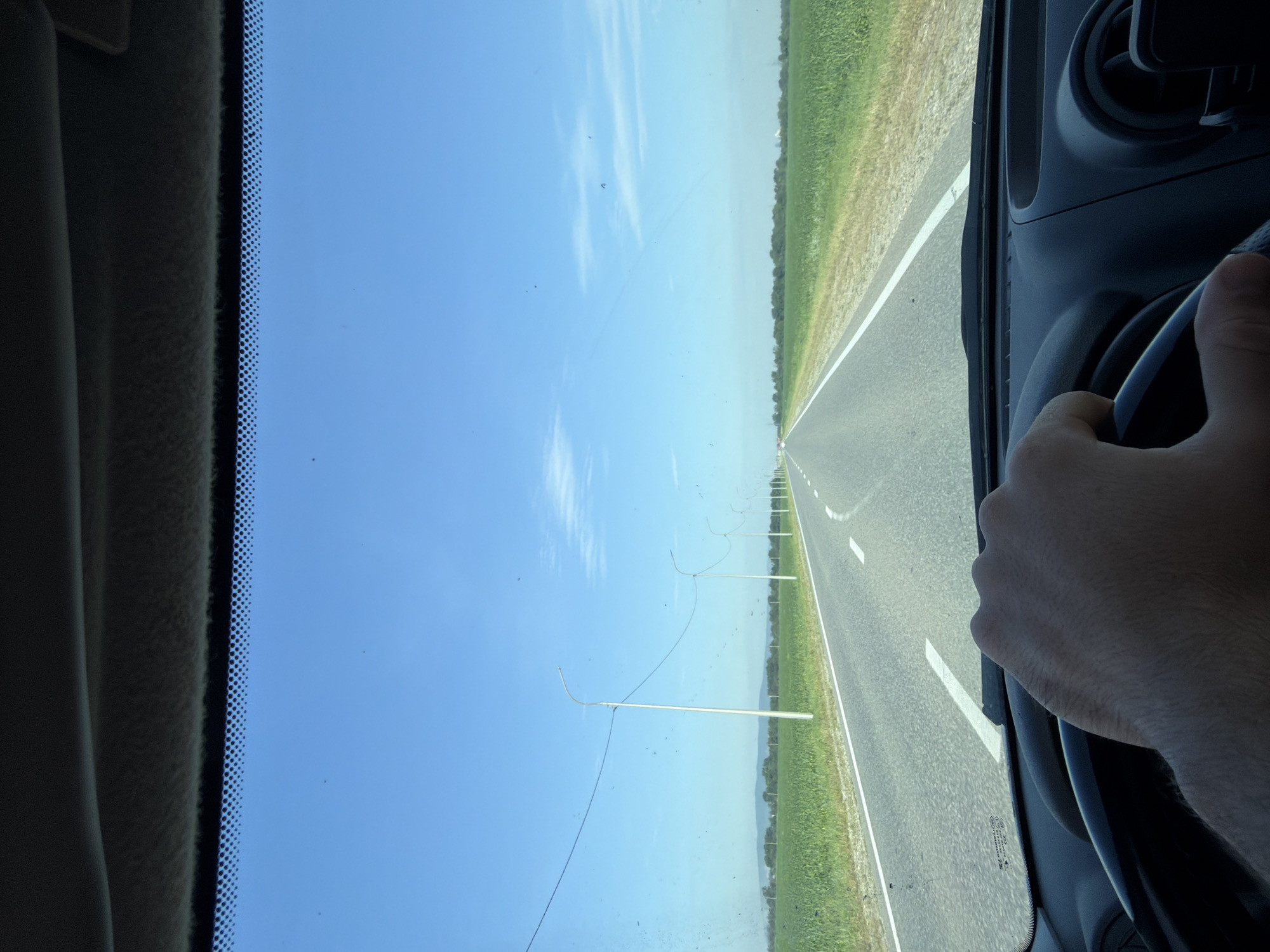 The image shows a first-person perspective from inside a vehicle, looking through the windshield. The vehicle is driving on a paved road that stretches into the distance, flanked by green fields on either side. The sky is a clear, bright blue with a few wispy clouds. The road has white lane markings. Streetlights are visible along the side of the road, spaced at intervals. In the foreground, a hand is visible gripping the steering wheel. The overall environment suggests a rural or suburban setting in Russia, possibly the outskirts of Mesker-Yurt. The image appears to be taken during the daytime.