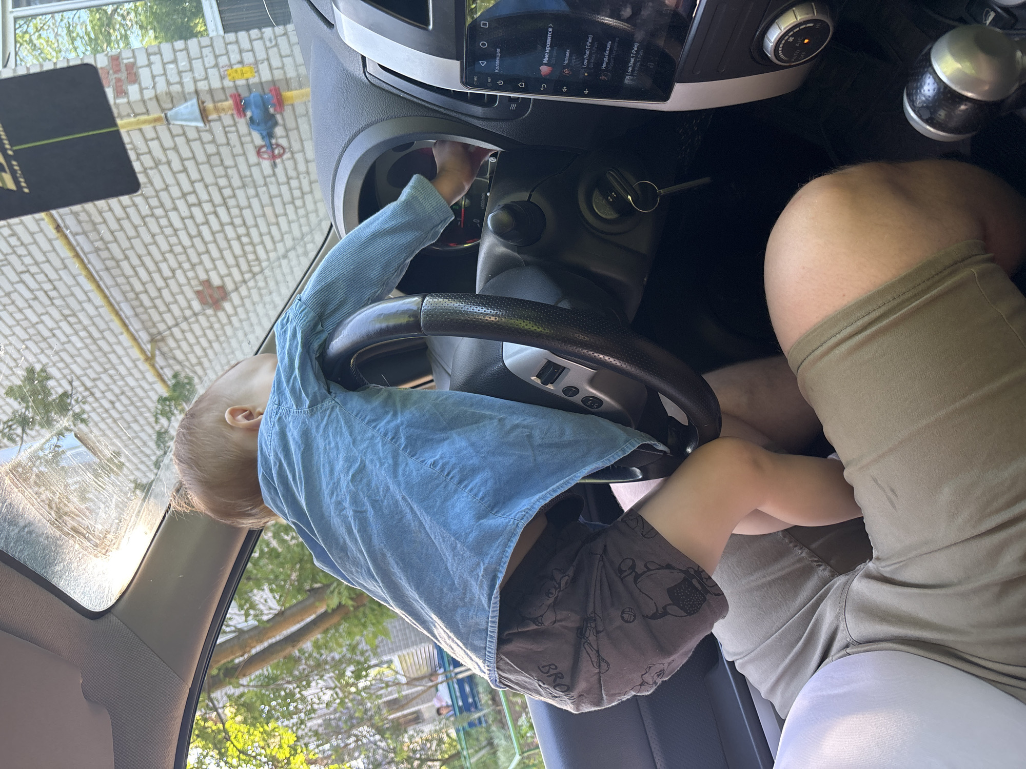 A young child wearing a blue shirt and patterned gray shorts is sitting on the lap of an adult wearing tan shorts inside a car. The child is reaching towards the car's dashboard and steering wheel. The car's interior is visible, including the steering wheel, dashboard, and infotainment screen displaying Russian text. Through the windshield, a view of a brick building with a yellow gas pipe and valve, trees, and a partially visible house can be seen. The image was taken in Горный, Russia.