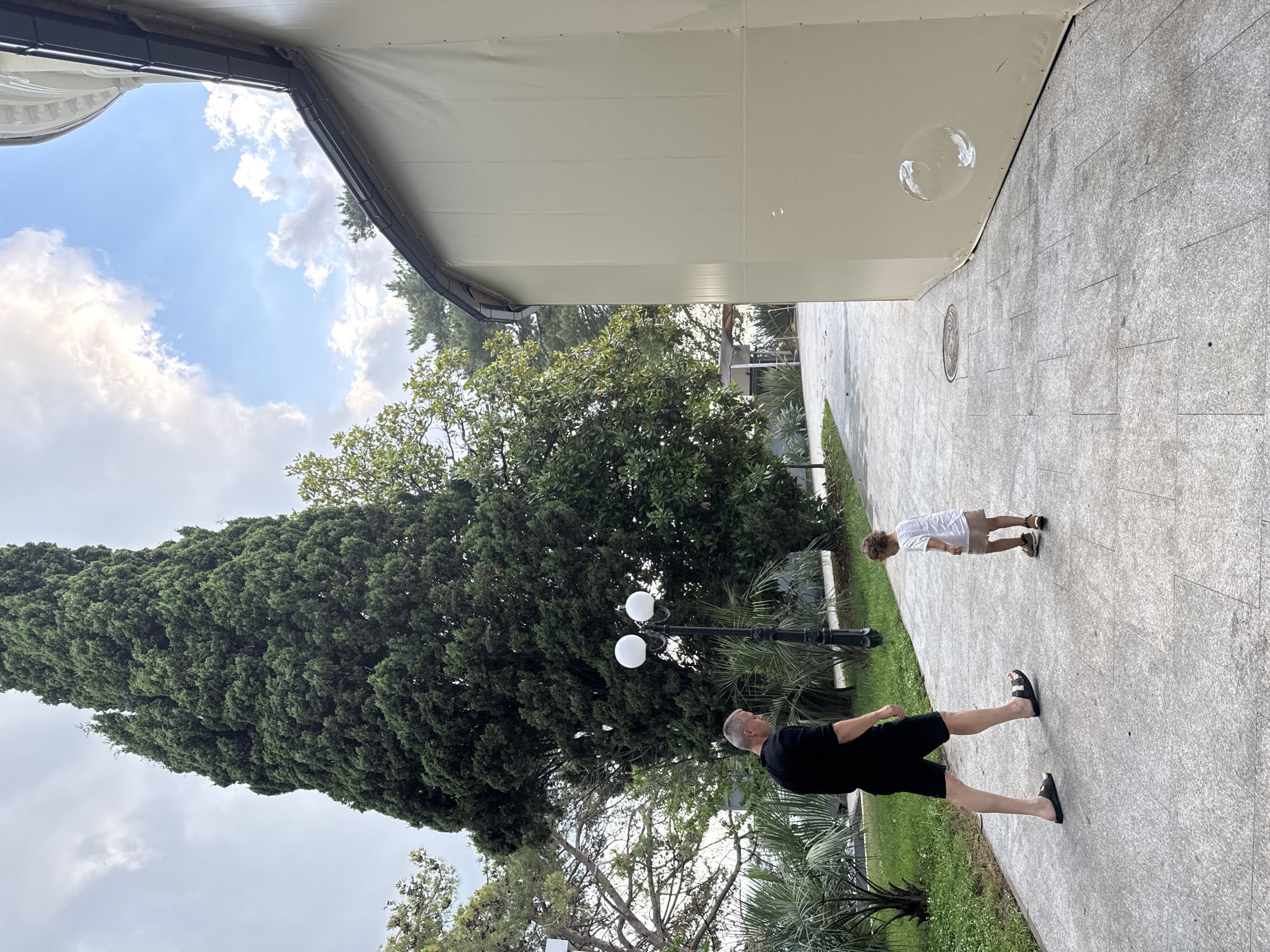 A man and a child are walking on a paved path in Sochi, Russia. The man is wearing a black t-shirt and black shorts, and the child is wearing a white t-shirt and a light brown skirt. A large evergreen tree is behind them, and a building with a tan awning is to their right. There are palm trees and other greenery around them. Two street lamps with round, white lights are also visible. The sky is partly cloudy. A bubble is floating near the building.