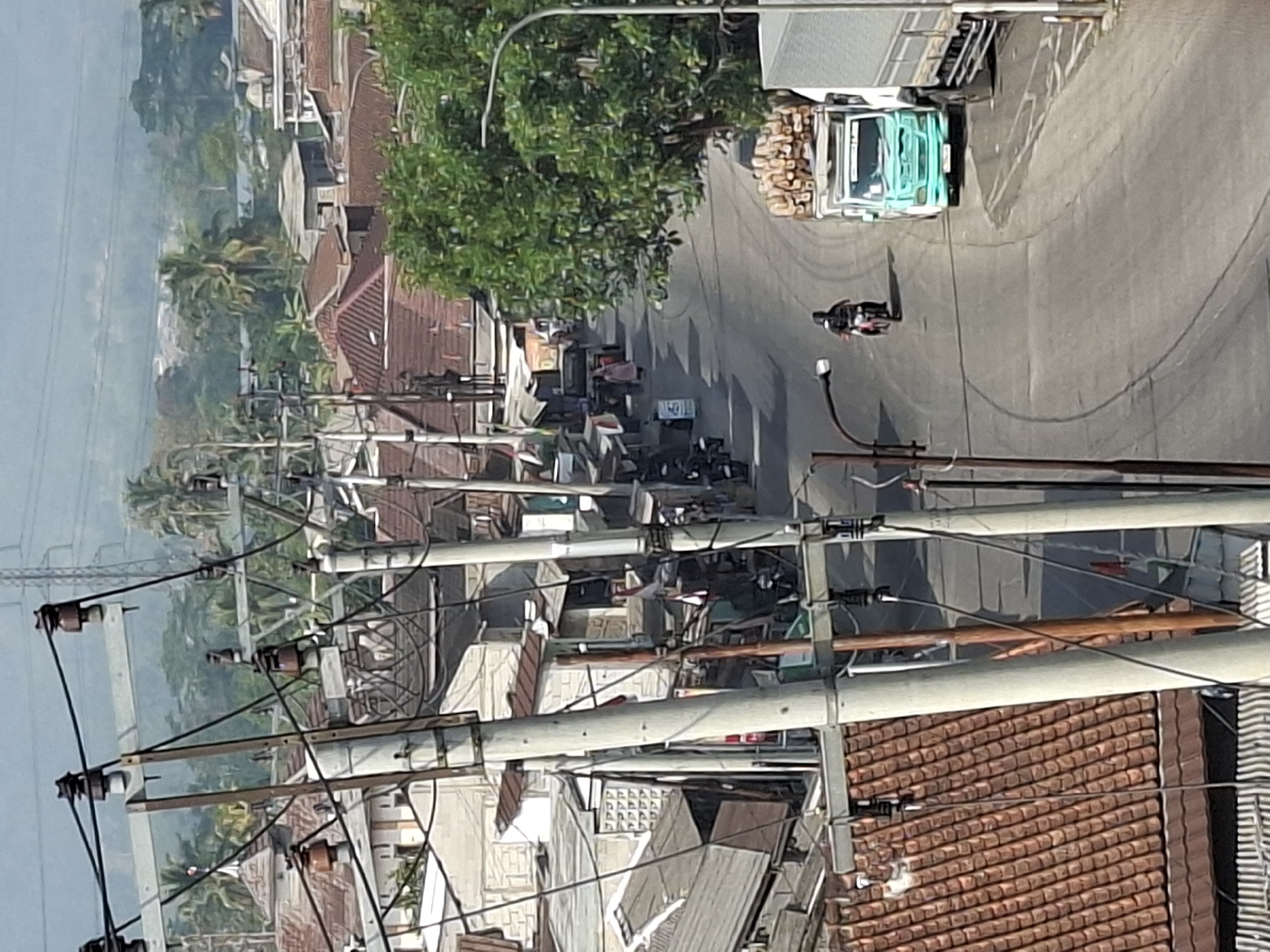 This is an outdoor, medium-distance, elevated shot of a street scene in Sukabumi, Indonesia. The foreground is dominated by several utility poles with electrical wires. The street curves through a densely populated area with houses and buildings featuring red-tiled roofs. In the middle ground, a person on a motorcycle is traveling down the street. To the right, a light blue truck is parked, loaded with what appears to be lumber or logs. Further down the street, a few people are walking. The background consists of lush green trees and hills, suggesting a somewhat rural or suburban setting on the outskirts of the city. The lighting suggests it is daytime, possibly late morning or early afternoon.