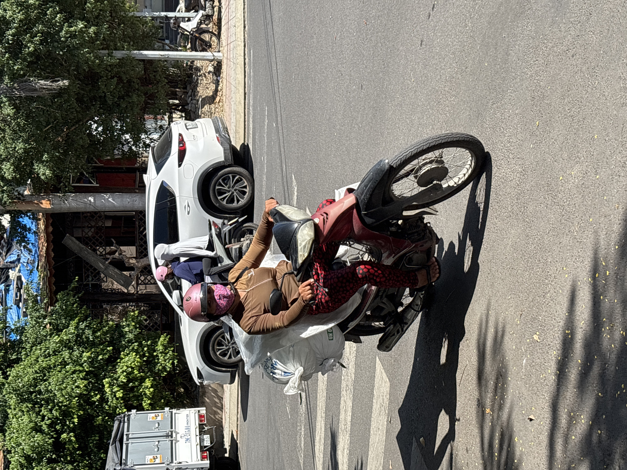 A woman wearing a pink helmet and a face mask rides a red and black scooter on a street in Nha Trang, Vietnam. She is carrying a large white bag of what appears to be recyclables strapped to the back of her scooter. She is wearing a brown long-sleeved shirt and red pants with a black polka dot pattern. Behind her, a white SUV and another scooter are visible. The scene is set on a sunny day, with shadows cast by trees and the scooter on the asphalt road. A pedestrian crossing is marked on the road in front of the scooter. In the background, there is a building with a partially visible roof covered in blue tarpaulin.