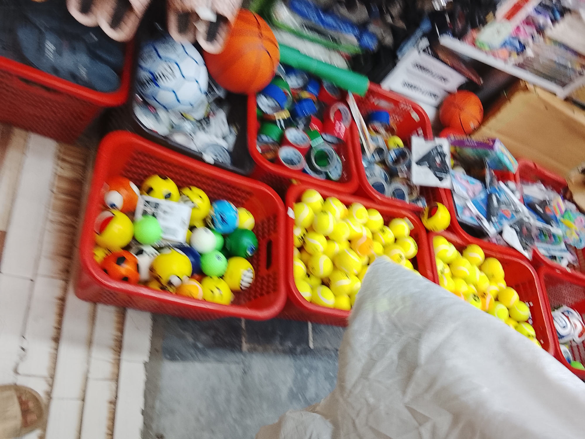 The image captures a display of toys in Gulgasht Colony, Pakistan. Several red plastic baskets are filled with various types of balls, including tennis balls, colorful toy balls, and miniature basketballs. A soccer ball is also visible. Other items such as toy guns, measuring tape, and children's footwear are scattered among the ball-filled baskets. The toys are displayed on what appears to be a concrete or tiled surface.