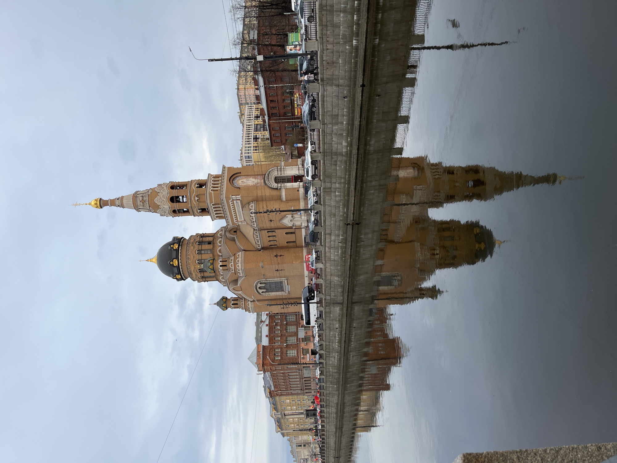 A cityscape is reflected in calm water under an overcast sky. The prominent structure is a large church building with a dark dome and a taller tower, both topped with golden spires. The reflection of the church is distinct in the water, mirrored along with adjacent buildings. A line of buildings and parked vehicles runs along the edge of the water, separating the reflected skyline from the actual buildings. The buildings consist of varying colors and architectural styles. Cars and some other vehicles can be seen parked on a narrow street or road between the building and the water. A stone embankment runs along the edge of the water on the bottom right corner of the frame. The absence of vibrant colors indicates a day with limited sunlight. There are no people visible in the scene. The location is an urban canal setting.