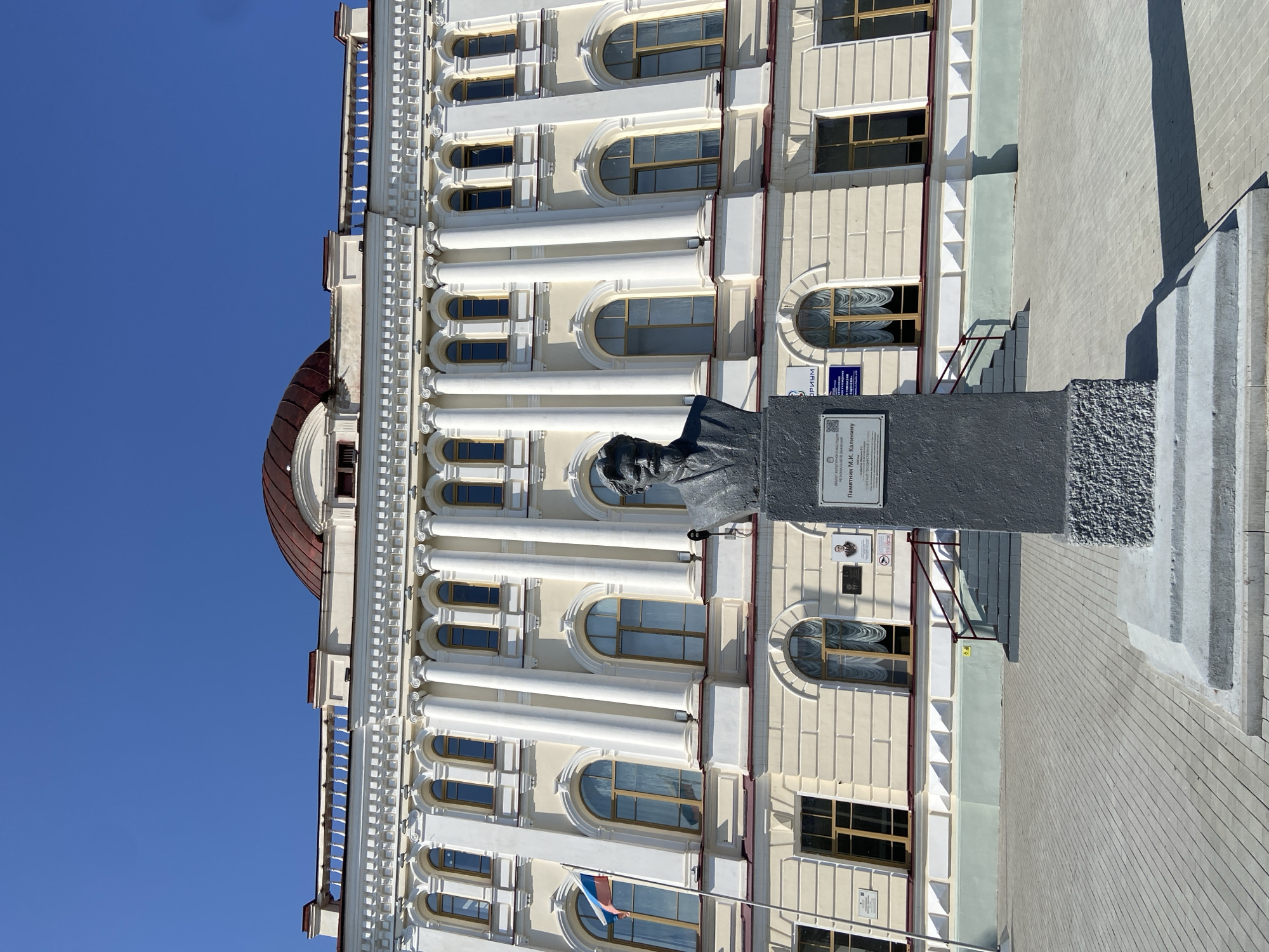 The image depicts the monument to M.I. Kalinin located in front of the building of the Blagoveshchensk State Pedagogical University, in the city of Blagoveshchensk, Russia. The building is a large, light-yellow structure with a classical facade, featuring rows of arched windows, white columns, and a red dome. A flag of Russia is visible on a flagpole to the left of the entrance. The bust of M.I. Kalinin, a prominent Soviet politician, stands on a grey pedestal in the foreground. The area in front of the building is paved with grey interlocking stones. The sky is clear and blue.