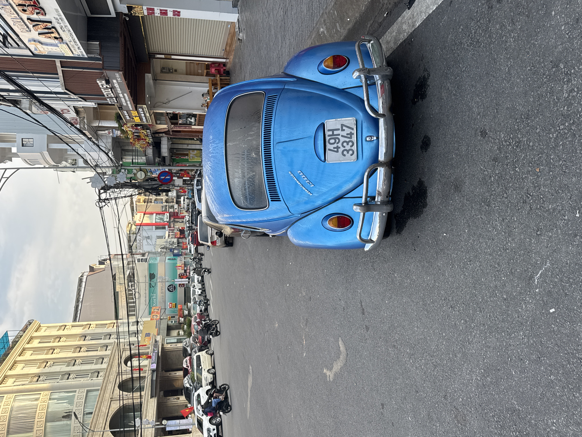 A candid daytime photograph captures a street scene in Da Lat, Vietnam. The primary subject is a vibrant blue, vintage Volkswagen Beetle, seen from the rear, parked on the right side of the asphalt road. The car features chrome bumpers and taillights, with a white license plate displaying "49H 3347". The word "VOLKSWAGEN" is faintly visible in script on its engine cover. The car and road show some dark, wet-looking patches, suggesting recent rain.
To the left and stretching into the background, the street is lined with multi-story buildings housing various businesses. Numerous motorbikes and several other cars are parked along the side of the road, reflecting typical urban activity in Vietnam. A network of utility cables crisscrosses overhead, adding to the urban landscape.
Visible storefront signs include "TEABREAK", "Kochi", "ANNA EYEGLASS", "NGỌC AN", "FEEL GOOD SPA", and "생하다 마사지" (Korean for "Fresh Massage"). Part of a Vietnamese sign reads "ƠNG ĐÃ LẠT", likely referring to local specialties of Da Lat. A red circular "no entry" sign is also visible. The sky is overcast, contributing to diffused lighting. The prevalence of motorbikes and the Vietnamese script firmly place the scene in Da Lat, Vietnam.