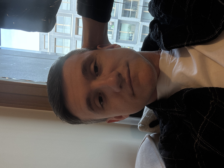 A male individual, appearing to be in his 20s or early 30s, is captured in a medium-shot selfie. He has short, dark hair and is looking directly at the camera with a neutral expression and a slight smirk. He is wearing a white t-shirt beneath a dark, possibly black or navy blue, quilted overshirt or jacket. His right arm is bent, with his hand positioned behind his head, and he appears to be seated or reclining on a light-colored sofa or armchair, with cushions visible to his left and behind his left shoulder. A plain, light-colored wall forms the background to his left. To his right, a large window reveals an urban environment. Through the window, a modern, multi-story building with a grid-like facade of numerous rectangular windows and light-colored panels is visible. Some sections of the building appear to have balconies with railings, and faint indications of greenery can be observed on some of these balconies. The sky is partially visible at the top of the window.
The scene is set indoors within a modern building in Warsaw, Poland. The outdoor background visible through the window depicts a contemporary urban district with high-rise architecture, consistent with newer residential or commercial developments within the city. A precise street address or specific neighborhood cannot be definitively determined from the visual evidence of the generic modern building.
