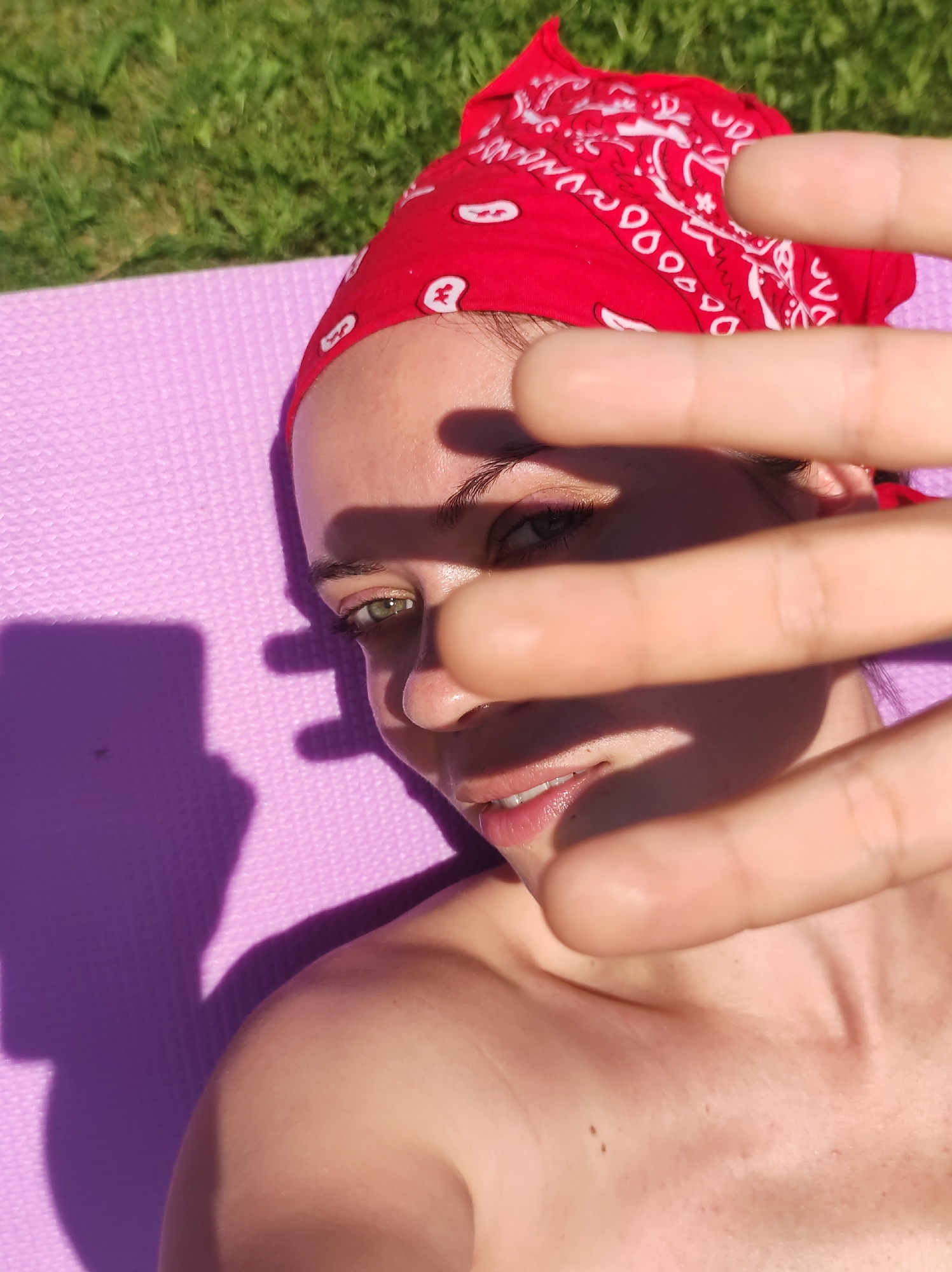 A fair-skinned woman with green eyes and a red bandana on her head, lying on a purple yoga mat on grass, shields her face with her hand from the sun. A shadow of a phone is visible on the mat, presumably used to take the picture. The location is not specified in the image and the city Фоки, Russia cannot be determined from the provided visual context.