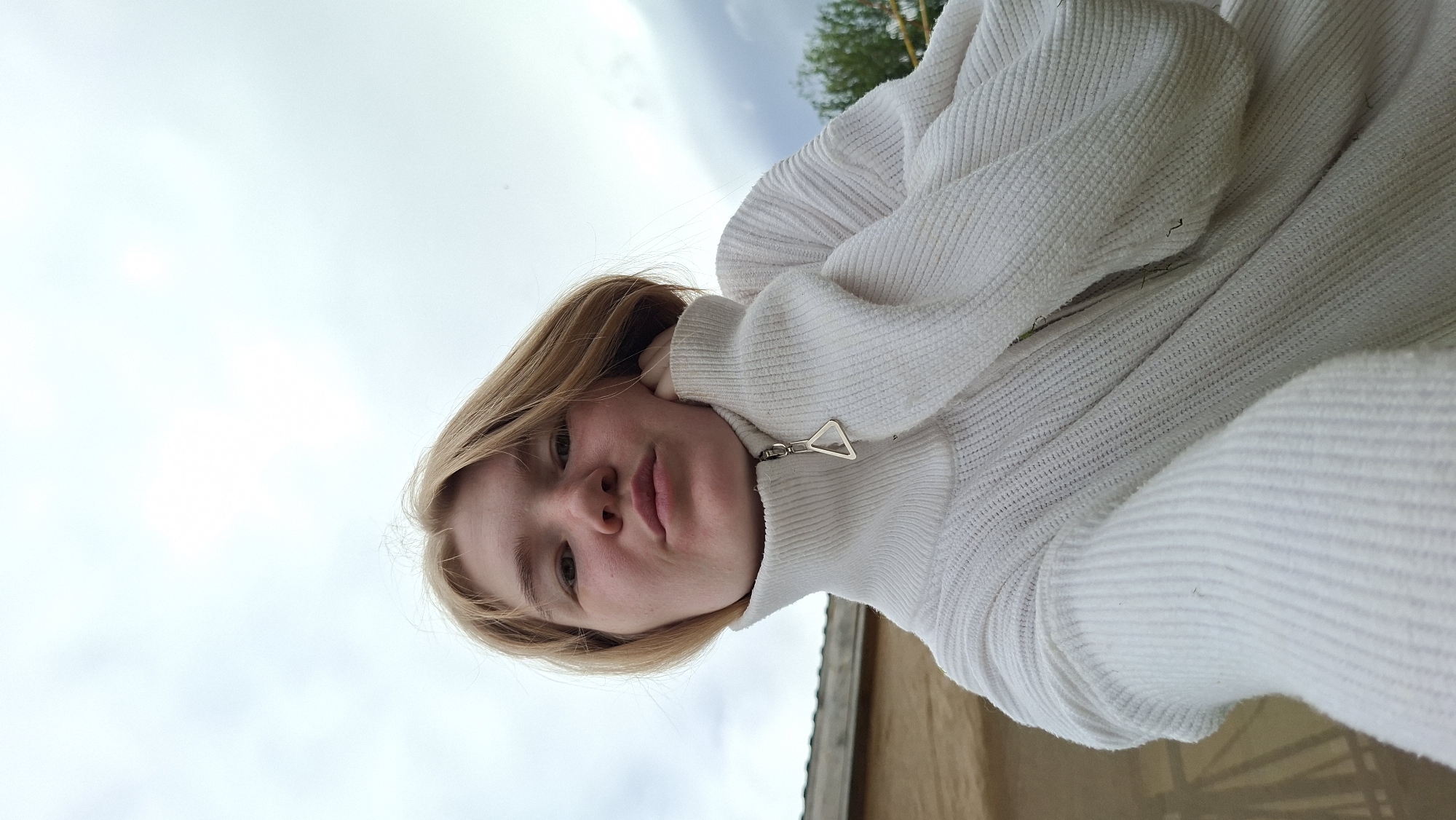 A young person with light blonde hair, wearing a white zippered turtleneck sweater, rests their chin on their hand. They are outdoors, with a cloudy sky visible in the background. To the left of the frame, a building with a light brown facade and a thatched roof is partially visible. The location is Big Bukor, Russia.