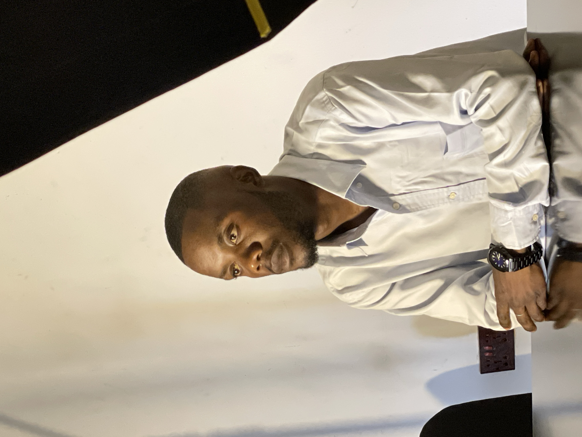 In Durumi, Nigeria, a dark-skinned man with a short haircut and a beard is seated at a table, his arms resting on its surface. He is wearing a light blue long-sleeved button-down shirt. A black wristwatch is visible on his left wrist. The man is looking to his left, away from the camera, with a neutral expression. The background consists of a textured white wall with a diagonal black panel on the right. A power outlet with multiple sockets is mounted on the wall behind the man. The table surface is reflective, showing a subtle reflection of the man and the background. The lighting is directional, creating shadows on the wall and highlighting the man's features and shirt.