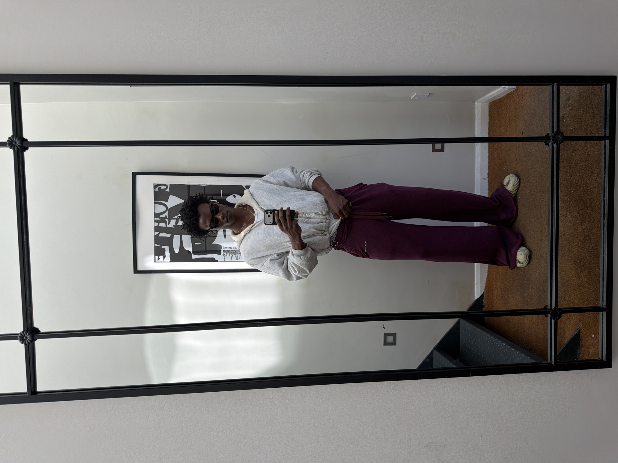 A person is taking a mirror selfie. The person is wearing sunglasses, a light-colored hooded sweatshirt, and burgundy-colored sweatpants. They are holding a cell phone in their right hand and are looking at the reflection in the mirror.
The mirror is a large, rectangular, multi-pane mirror with a black frame. A piece of art hangs on the wall. The background shows part of a staircase and a wall with a light switch or outlet. Due to the lack of external clues in the environment, the city of London, United Kingdom cannot be identified with confidence.