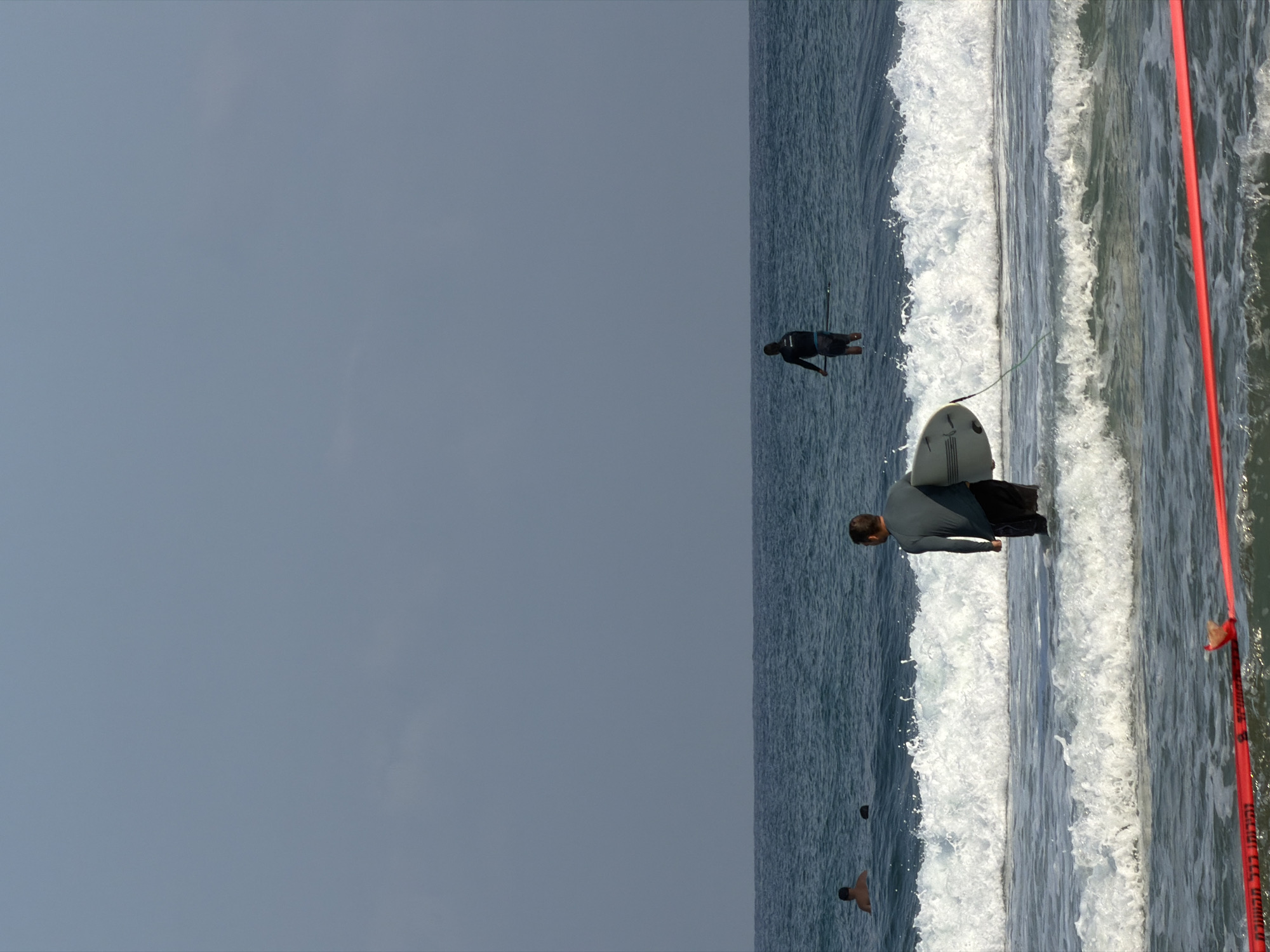An outdoor, full-shot, eye-level, vertical, medium-long shot depicts a body of water and a hazy, pale blue sky. In the foreground, a person in a dark-colored, long-sleeved rashguard and dark shorts stands in the ocean, holding a light-colored surfboard. The person is facing away from the camera and towards the right of the frame. Waves are breaking around the person. Further out in the water, another person, wearing a dark long-sleeved top and dark shorts, stands on a paddleboard, holding a paddle, and facing away from the camera. In the background, two more individuals are visible in the water, appearing to swim. A red warning tape is stretched across the bottom of the frame, partially submerged in the water. The scene is set on a beach in Bat Yam, Israel.