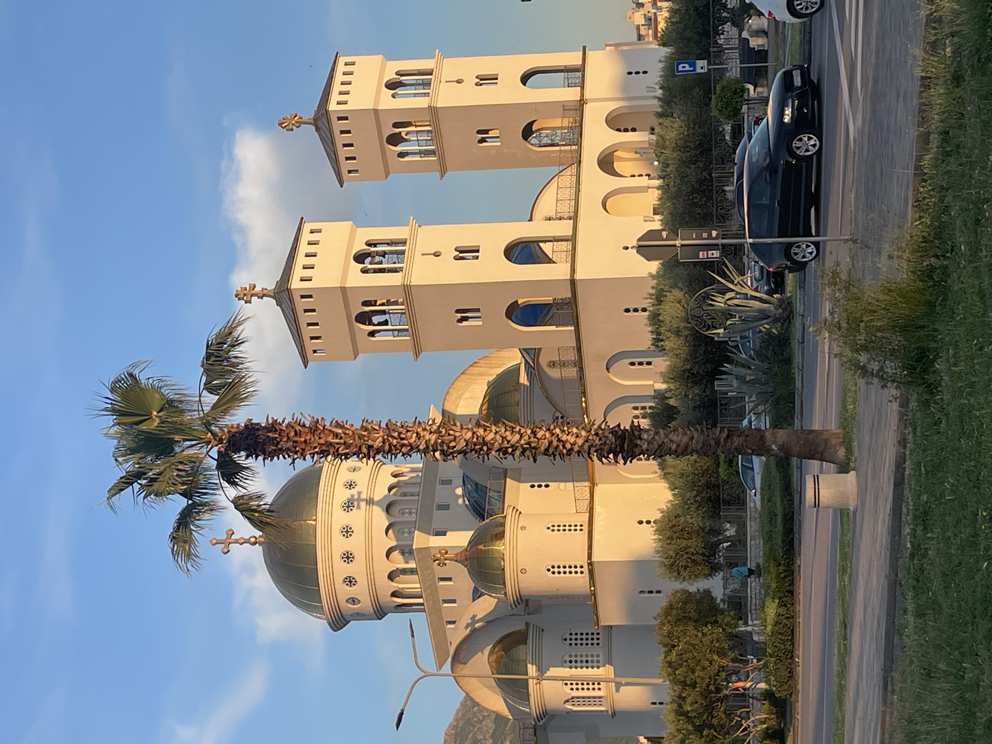 The image shows the Church of St. John Vladimir in Bar, Montenegro, from a street-level perspective. The church is predominantly white with golden domes and crosses on top. Two bell towers stand tall at the front of the church. A tall palm tree stands between the viewer and the church, partially obscuring the building. To the right of the palm tree and church, a road is visible with cars parked along the side, a car driving down the road and a parking sign post. Bushes and green vegetation run alongside the road. The sky is blue with some light clouds. The image is taken in daylight.