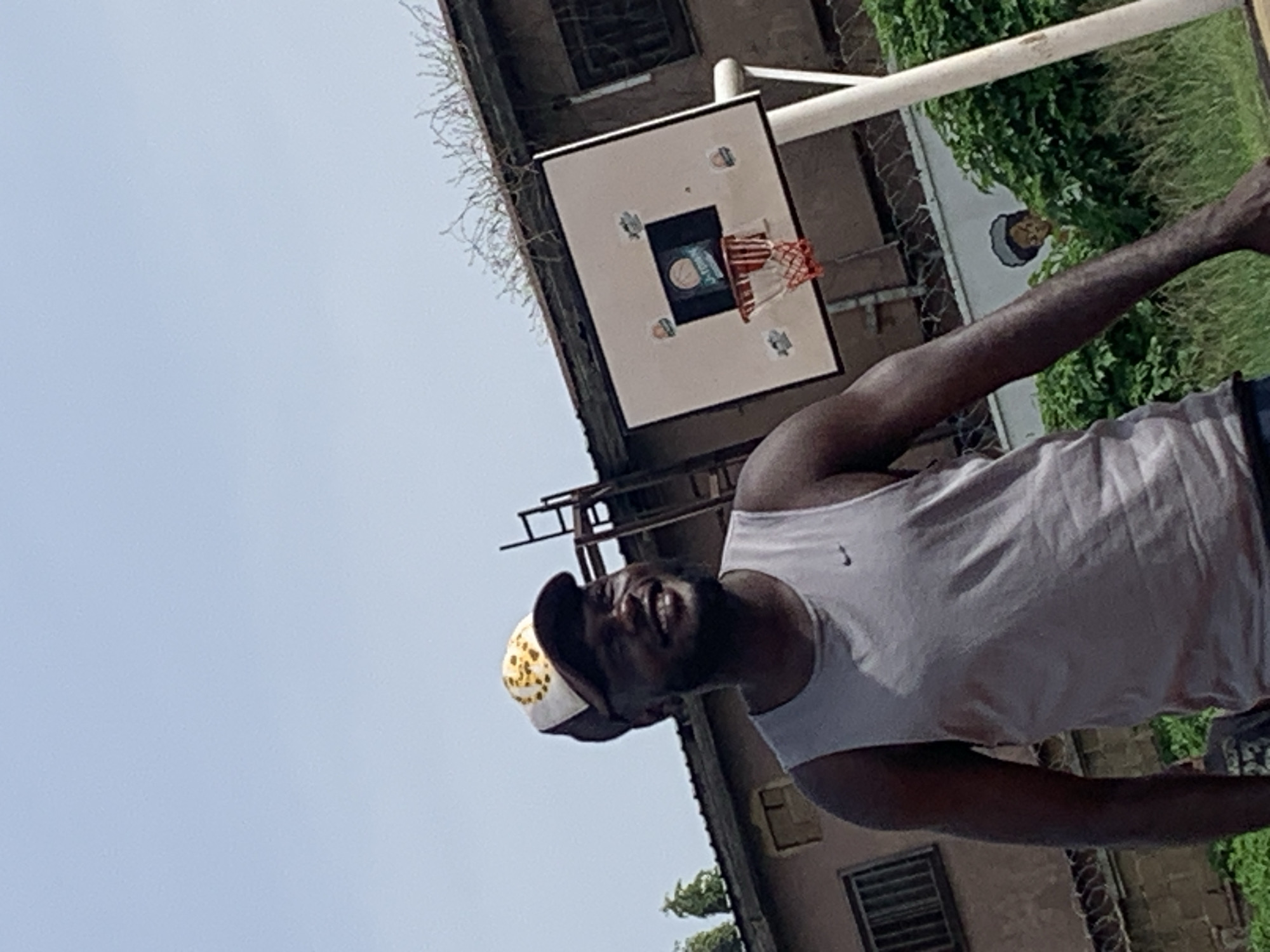 An outdoor shot, presumably in Gura Top, Nigeria, features a man standing in front of a basketball hoop. The man, wearing a gray tank top and a baseball cap, is smiling and looking upwards and to his right. The basketball hoop has a white backboard with several stickers and a red net. Behind the hoop, a brown building with barred windows is visible. Lush green foliage and a wire fence with barbed wire line the ground. The sky is clear and blue.