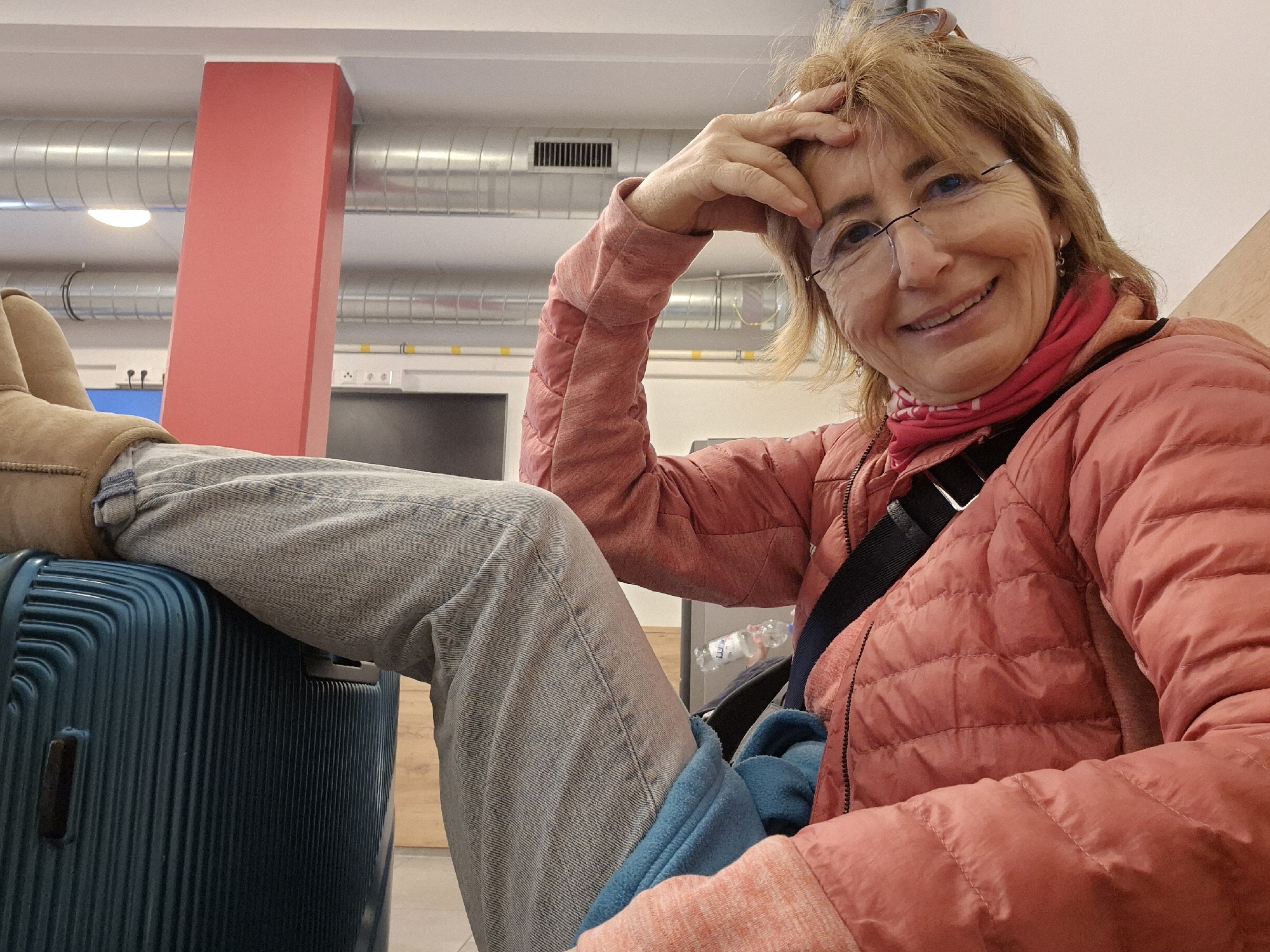 A middle-aged woman is the primary subject of the image. She has fair skin, short blonde/red hair, and wears glasses. She is smiling at the camera and is wearing a pink quilted jacket and a patterned scarf. A black strap crosses her body, likely from a purse or bag. Her legs are stretched out, one resting on top of a blue suitcase. She wears grey jeans and light brown boots. Her right hand is touching her forehead.
The setting appears to be an indoor waiting area, possibly an airport or train station. A red pillar is visible in the background, along with metal ventilation ducts near the ceiling. A power outlet is attached to the wall. A water bottle is situated near her legs.
While there is no explicit evidence in the image to confirm that it was taken in Vienna, Austria, the architectural and infrastructural elements do not contradict this. Without additional information such as signage, identifying features, or contextual clues, this cannot be verified.