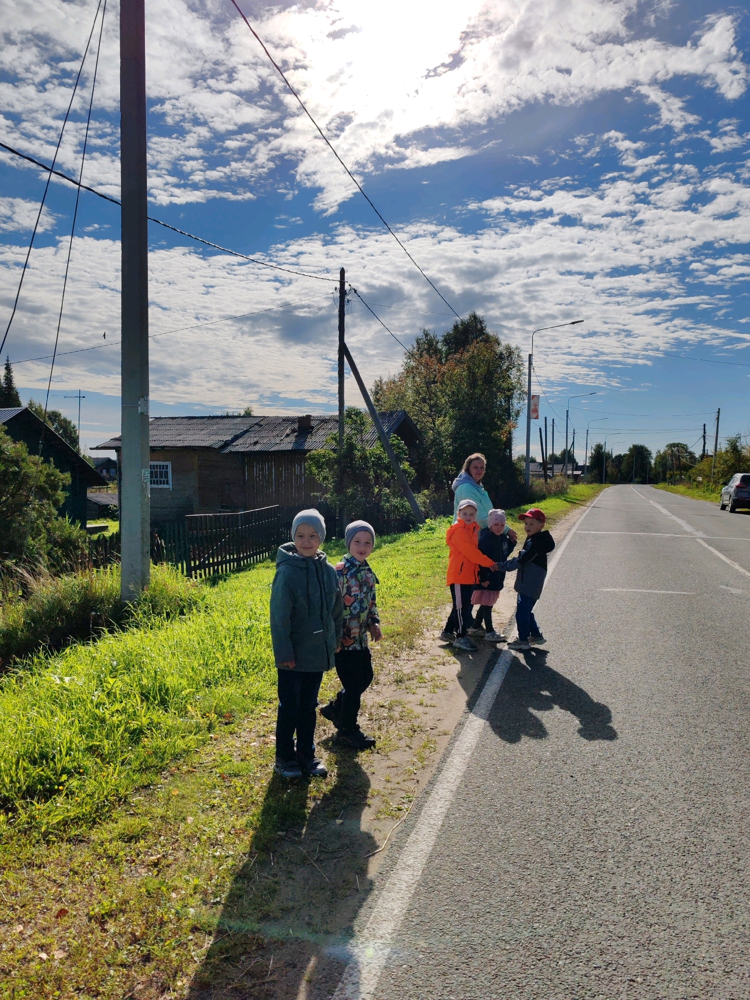 In Чухлэм, Russia, on a sunny day, a group of five children and one adult stand on the grassy shoulder of a paved road. The adult, wearing a light blue hooded jacket, is positioned behind the children, who are all facing the road. Three of the children are wearing hats: one in a gray beanie and two in pink and red caps. One child in an orange jacket is holding hands with another child in a pink skirt and jacket. A child in a gray jacket stands closest to the foreground. The road appears to be a two-lane highway with a white line marking the edge. In the background, a wooden house with a dark roof and a white-windowed facade is visible, surrounded by trees and other structures. Utility poles and wires crisscross the bright blue sky, which is dotted with white, fluffy clouds. The sun is high in the sky, casting long shadows of the people onto the road.