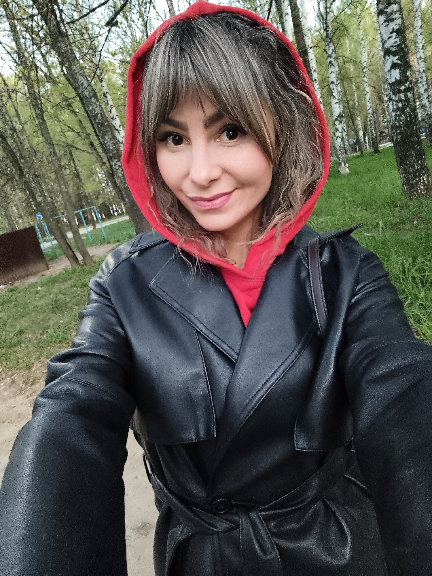 A woman with bangs and curly hair, wearing a red hoodie under a black leather trench coat, takes a selfie. The background shows a park with birch trees and green grass. In the distance, a small playground with blue and green elements is visible. The location is likely Chaikovsky, Russia, based on the photographer's known location.