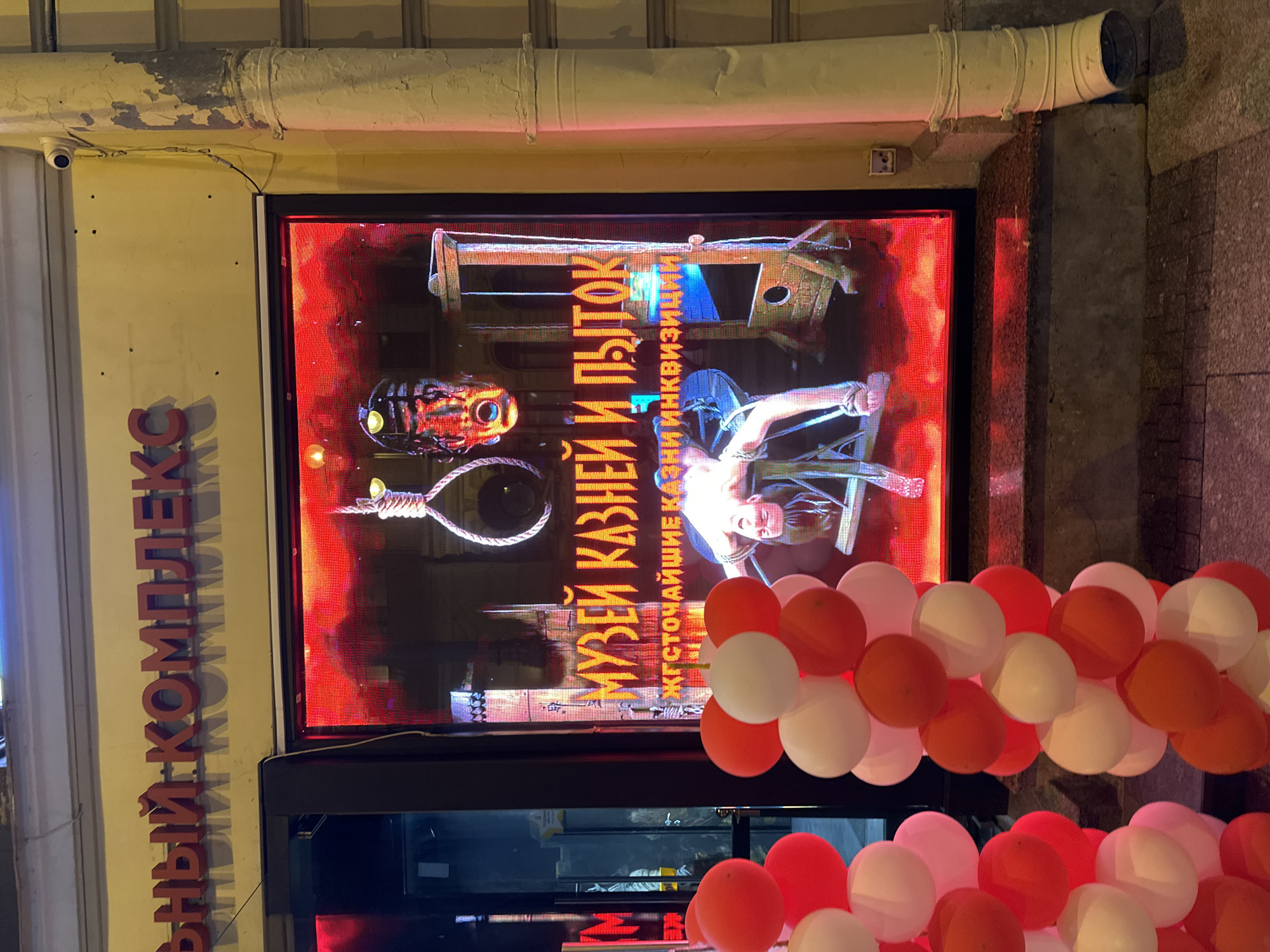 Here is a forensic description of the image:
**Location:** The image shows a street scene in an unidentified city in Russia, based on the Cyrillic script on the signage.
**Objects:** The primary focus is a large, brightly lit display screen in a window. The display shows a graphic image related to torture and executions. Key elements on the screen include:
* A guillotine.
* A torture mask.
* A noose.
* A person strapped to a chair.
* Text in Russian, possibly advertising a "Museum of Executions and Torture."
In the foreground, there are several bunches of balloons, predominantly in red, white, and orange. The balloons are partially obscuring the bottom of the display screen. The building facade is visible, painted yellow with a white pipe running along it. A security camera is mounted on the facade.
**Text/Signage:** There is vertical text in Cyrillic lettering on the building facade to the left of the display screen. The text on the display is in Russian.
**People/Subjects:** The image on the display screen depicts a person in a potentially vulnerable state, strapped to a chair. The graphic is part of the museum's promotional material.
**Environment:** The lighting suggests an evening or nighttime setting. The architecture of the building appears older. The environment has an air of a slightly run-down city street.
