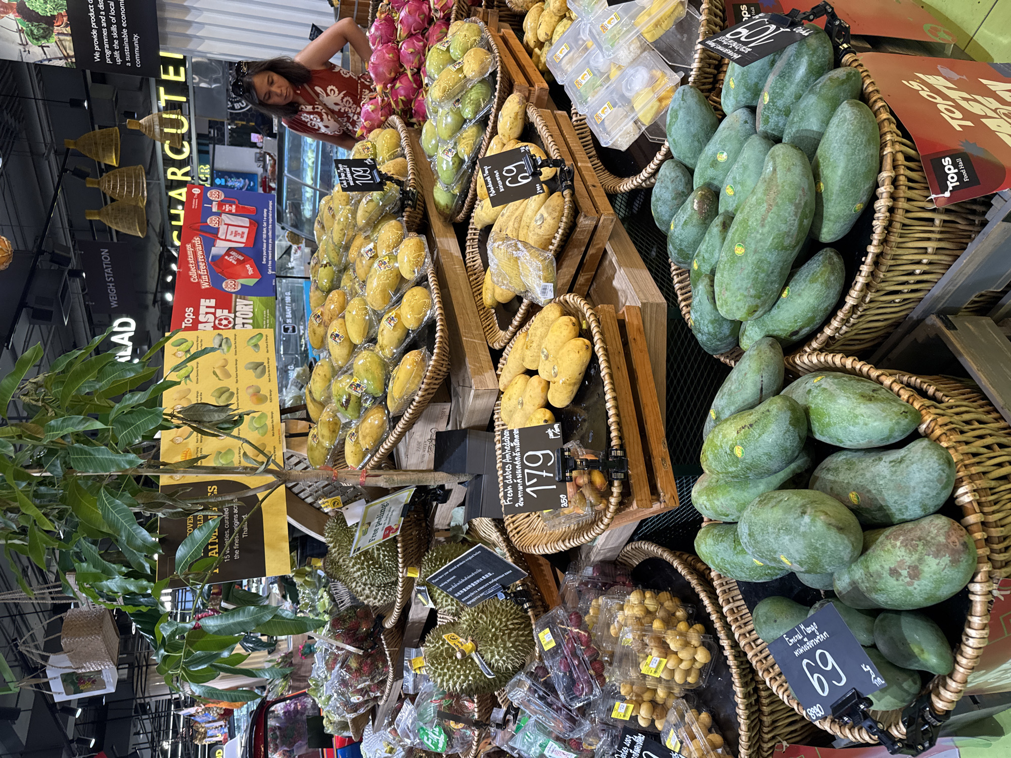 The image shows a produce section of a grocery store, likely Tops Market, in Koh Samui District, Thailand. Several varieties of fruits are displayed in woven baskets and plastic containers, including mangoes, durians, rambutan, and dragon fruit. A woman in a red patterned top is visible in the background, looking at the fruit display. Signs in Thai and English advertise prices and promotions, such as "Taste of History" and "Collect stamps. Win free rewards." The overall environment is a well-lit indoor market with a focus on fresh produce.