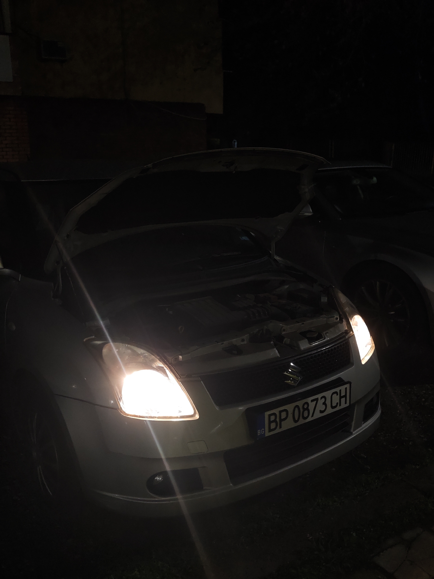 A nighttime scene depicts a white Suzuki Swift car with its hood open and headlights illuminated. The license plate reads "BP 0873 CH," indicating registration in Bulgaria. The car is parked next to another vehicle, partially visible. The location appears to be an urban area, judging by the building visible in the background, though the overall darkness obscures specific details about the surroundings. No individuals are visible in the frame. The image appears to be taken in Kozloduy, Bulgaria, judging by the Bulgarian license plate.