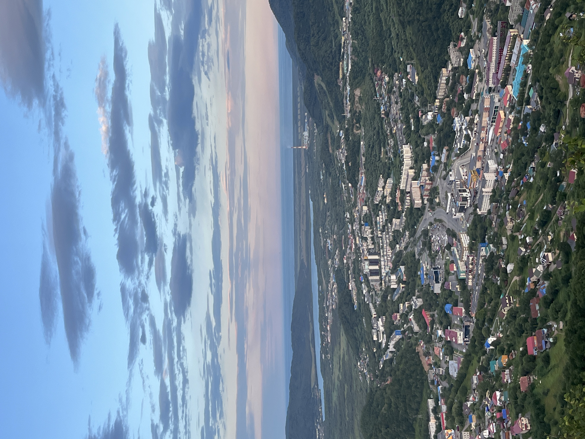 This is a high-angle, vertical shot overlooking the city of Petropavlovsk-Kamchatsky, Russia, at dusk. The sky is a blend of pale blue and soft pinks, with scattered clouds. In the foreground, a densely packed residential area with a mix of multi-story apartment buildings and smaller houses with colorful roofs blankets a lush green hillside. A winding road cuts through the urban sprawl, lined with more buildings and vehicles.
Further back, the city expands into a flatter valley, featuring a large body of water, possibly a bay or lake. Beyond the water, a power plant with a tall, striped smokestack is visible, emitting a thin plume of smoke. Rolling, tree-covered hills form the backdrop, meeting the distant horizon where the ocean meets the sky. The overall impression is of a compact city nestled within a verdant and coastal landscape, observed from a high vantage point as daylight fades.