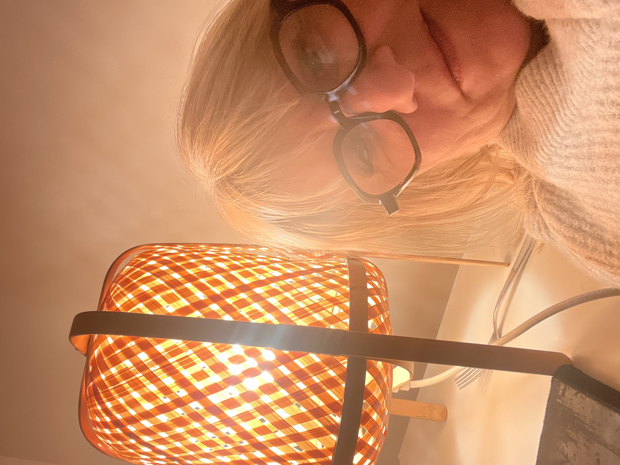 A selfie-style photograph looking down at a lit lamp with a woven orange-toned shade from a person with blond hair and glasses. The person is partially visible in the upper portion of the frame and is wearing a light-colored sweater. The lamp is the primary focus, with the light source appearing to be bright and centered within the shade. The background appears to be a plain wall, with a power cord and wall outlet visible. The photograph location is Chambœuf, France.
