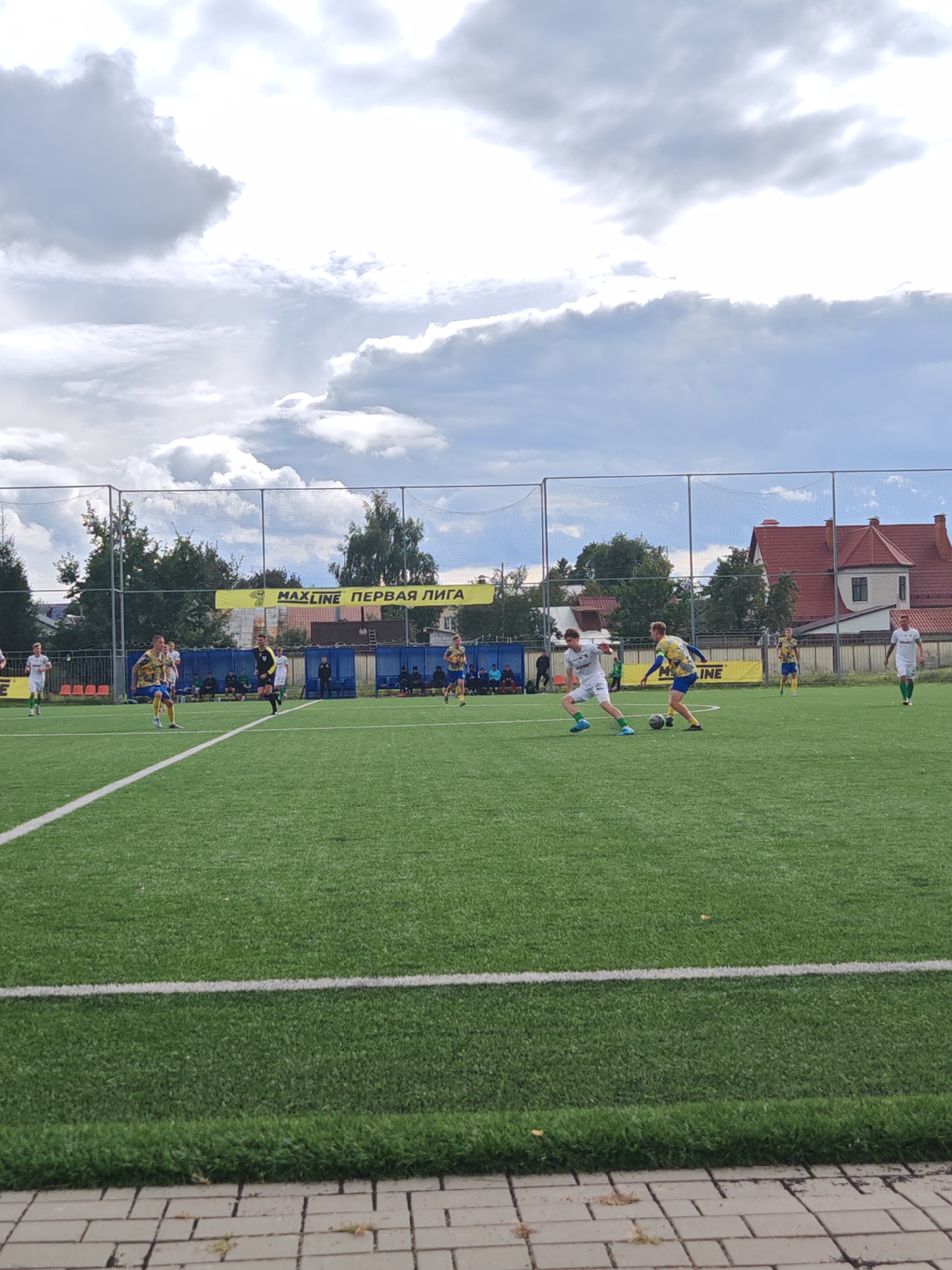The image shows a football match on an artificial turf field. Several male athletes in team uniforms are engaged in play. One player in a white jersey with green trim is dribbling the ball, while another player in a blue and yellow jersey is attempting to intercept. A referee in a black uniform is observing the play. In the background, a yellow banner with "MAXLINE ПЕРВАЯ ЛИГА" is displayed. Several spectators are seated on bleachers and standing behind a fence. The scene is set outdoors under a cloudy sky, with trees and buildings visible in the distance. The location is identified as Homyel, Belarus.