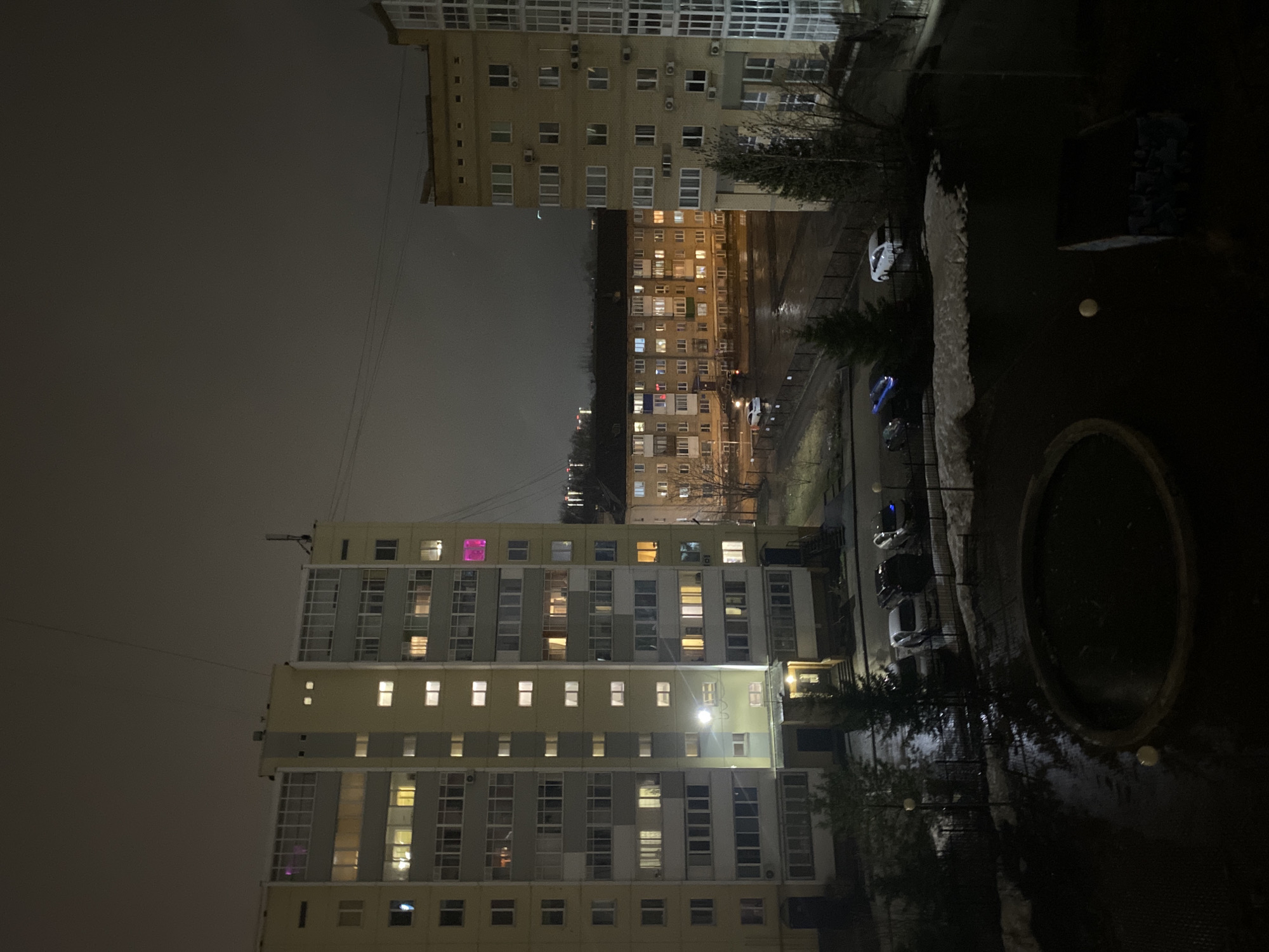 The image shows a nighttime scene in Perm, Russia. Two apartment buildings are visible, illuminated by interior lights. A street with parked cars runs between them. The foreground contains a dark, circular object, likely a fountain or planter. The sky is overcast.
