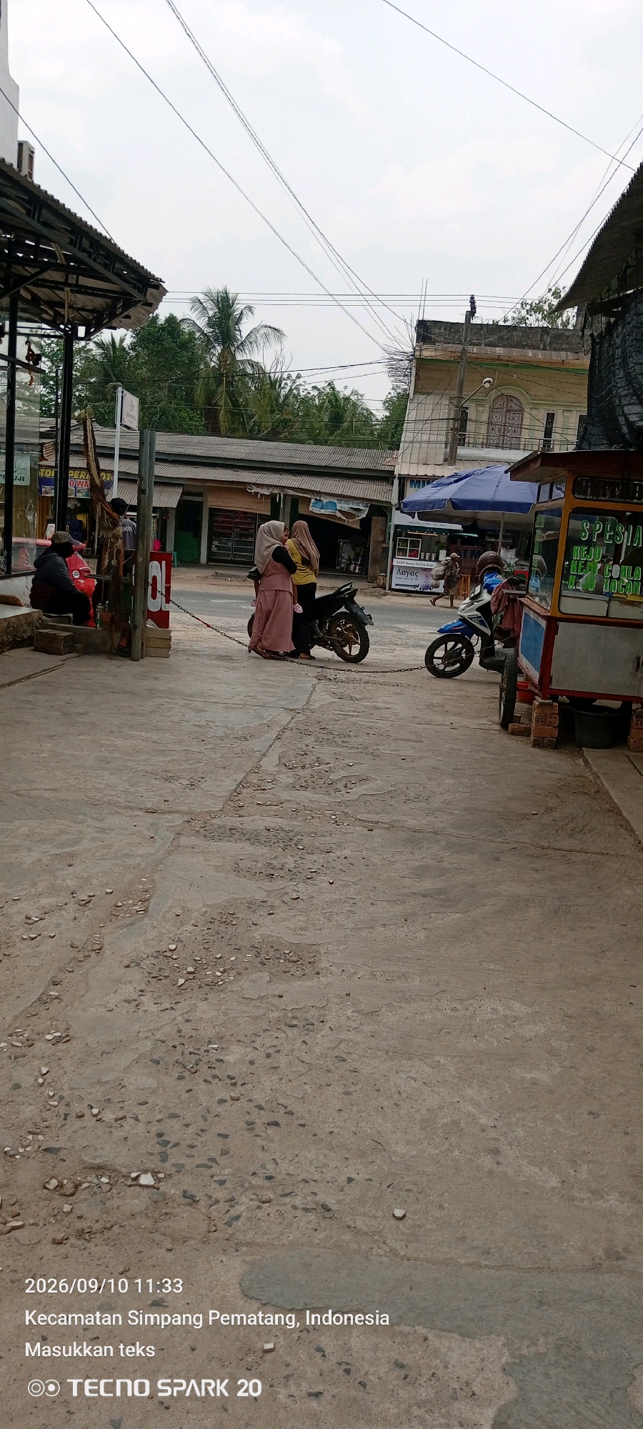 This image captures a street scene in Simpang Pematang, Indonesia. In the foreground, a concrete road surface shows signs of wear and tear, with scattered gravel and cracks. On the left, a man wearing a dark cap and jacket sits on a wooden crate. To his right, a metal chain extends across the pavement, linking to a motorcycle parked further down the street. Two women, both wearing hijabs and modest clothing, stand near the motorcycle. One woman is dressed in pink, while the other wears a yellow top and a brown hijab.
In the middle ground, several storefronts with corrugated metal roofs line the street. A blue umbrella is set up in front of one of the shops. Beyond the shops, a multi-story building with arched windows is visible. A street vendor's cart, painted red and yellow, is positioned on the right side of the image. The cart has text on its side, including the word "SPESIAL".
The background is dominated by lush green trees, including palm trees, and a cloudy sky. Power lines are strung across the sky, adding to the urban atmosphere. The timestamp on the image indicates it was taken on 2026/09/10 at 11:33. The location is identified as Kecamatan Simpang Pematang, Indonesia.