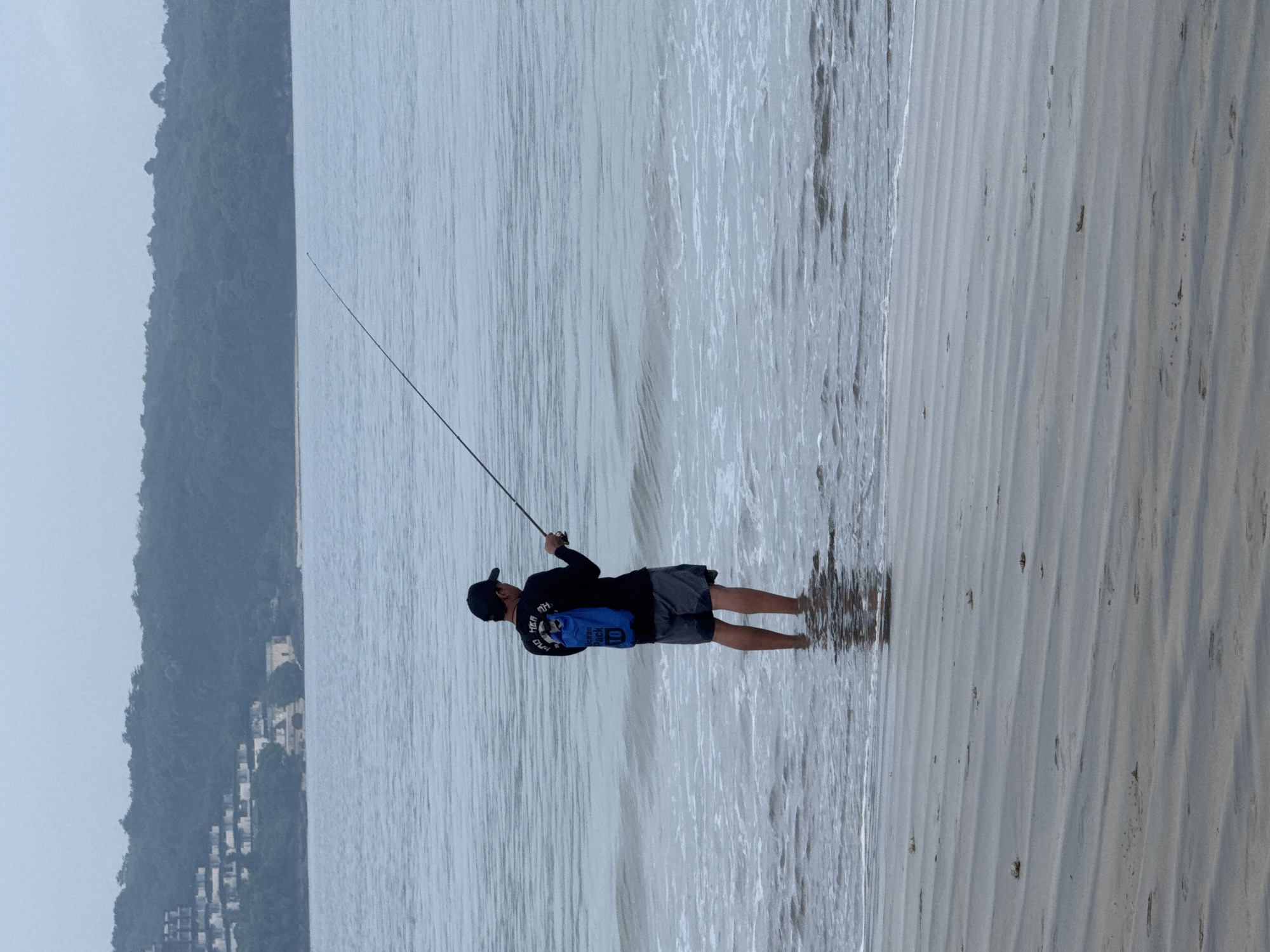 A man is fishing at Jimbaran beach in Jimbaran, Indonesia. The man stands in the water with a fishing rod in his hands. He is wearing a dark long-sleeved shirt, dark shorts, and a dark baseball cap. Behind him, the ocean stretches out towards the horizon. To the left, a wooded area is visible with buildings nestled amongst the trees. The sky appears overcast. The beach sand shows ripple patterns from the receding tide.