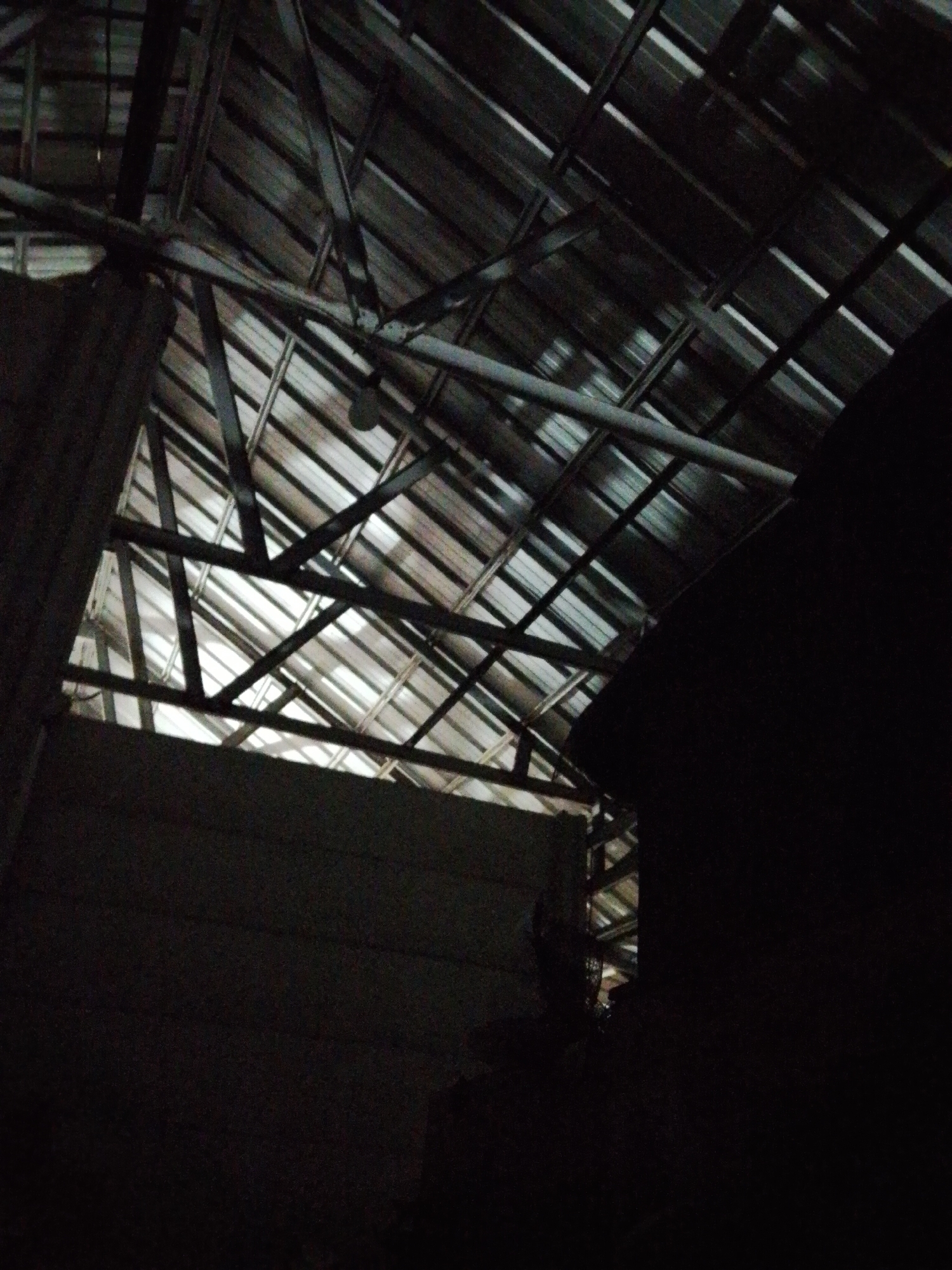 The image shows the interior of a building in Bukit Ketok, Indonesia. The structure appears to be an industrial or agricultural building, judging by the metal roofing and framework. Light is visible through gaps in the roof. The scene is dimly lit, with areas of heavy shadow in the foreground. Walls are visible on the left and right.