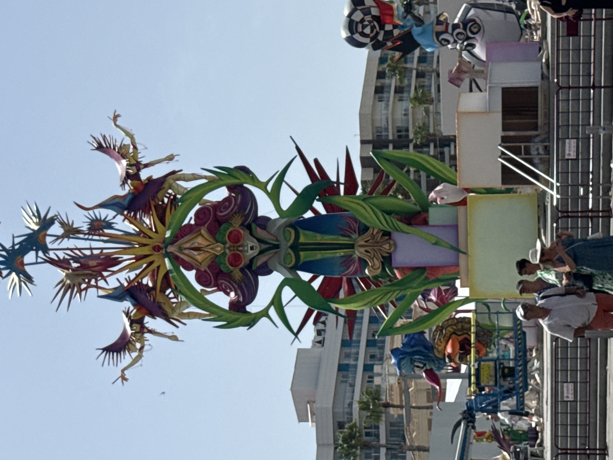 A large, elaborate, and colorful papier-mâché sculpture stands tall against a clear blue sky, dominating the center of the frame. The sculpture depicts a stylized, mythical figure with outstretched arms, adorned with vibrant purple, green, yellow, and red hues. The figure has insect-like wings and appears to be in mid-flight or dance. Surrounding the central figure are other elements of the sculpture, including spiky, star-like protrusions and swirling decorative motifs.
In the background, modern buildings with balconies and palm trees suggest an urban coastal setting. The sculpture is situated outdoors, possibly in a public square or during a festival, as evidenced by a metal fence in the foreground. Several people are visible in the lower portion of the image, walking or standing near the fence. One man in the foreground wears a white shirt and orange shorts, facing away from the camera. Behind him, three other individuals are clustered together. Further back, a blue lift is partially visible, with people working on or near it, suggesting ongoing setup or maintenance of the display. The overall scene is brightly lit by the sun.